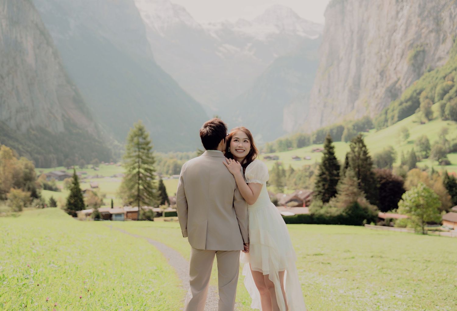Bride smiling over her shoulder while groom embraces her on a green meadow with Alpine mountains behind.