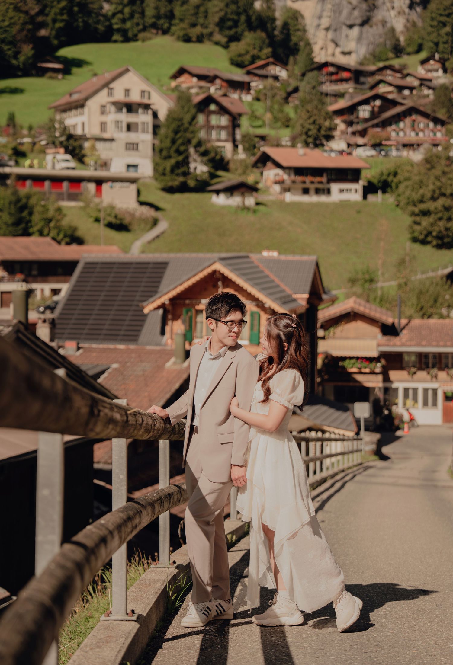Couple walking hand in hand along a wooden fence with a scenic Swiss village and cliffs behind them.