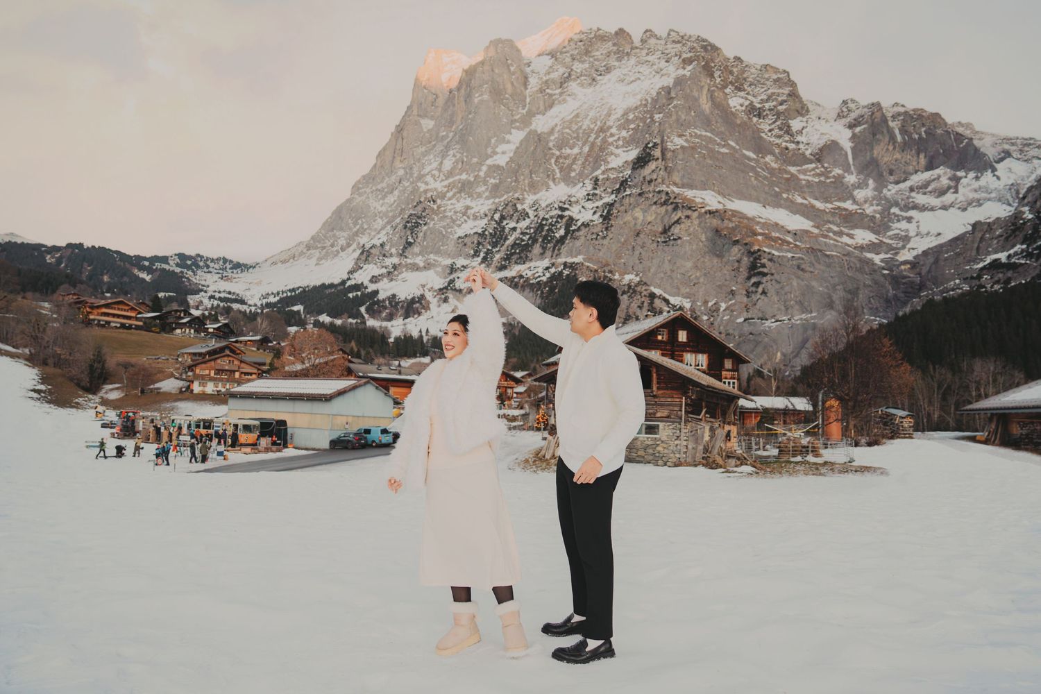 Joyful couple in white outfits dancing on snow with scenic alpine village and mountain behind them.