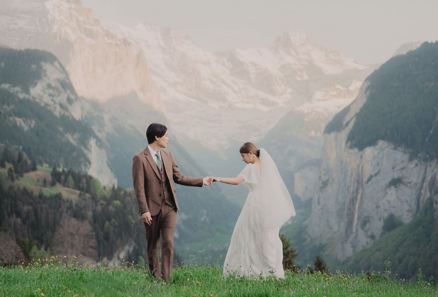 Bride and groom holding hands facing each other in a stunning alpine meadow with misty Swiss mountain backdrop.