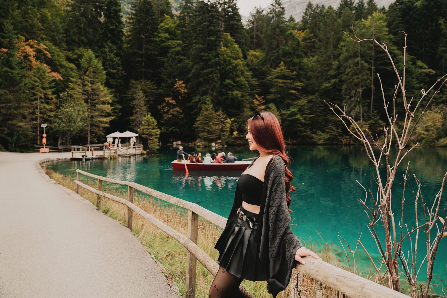 Woman with red hair stands by a wooden fence overlooking a turquoise alpine lake with boats and forest backdrop.