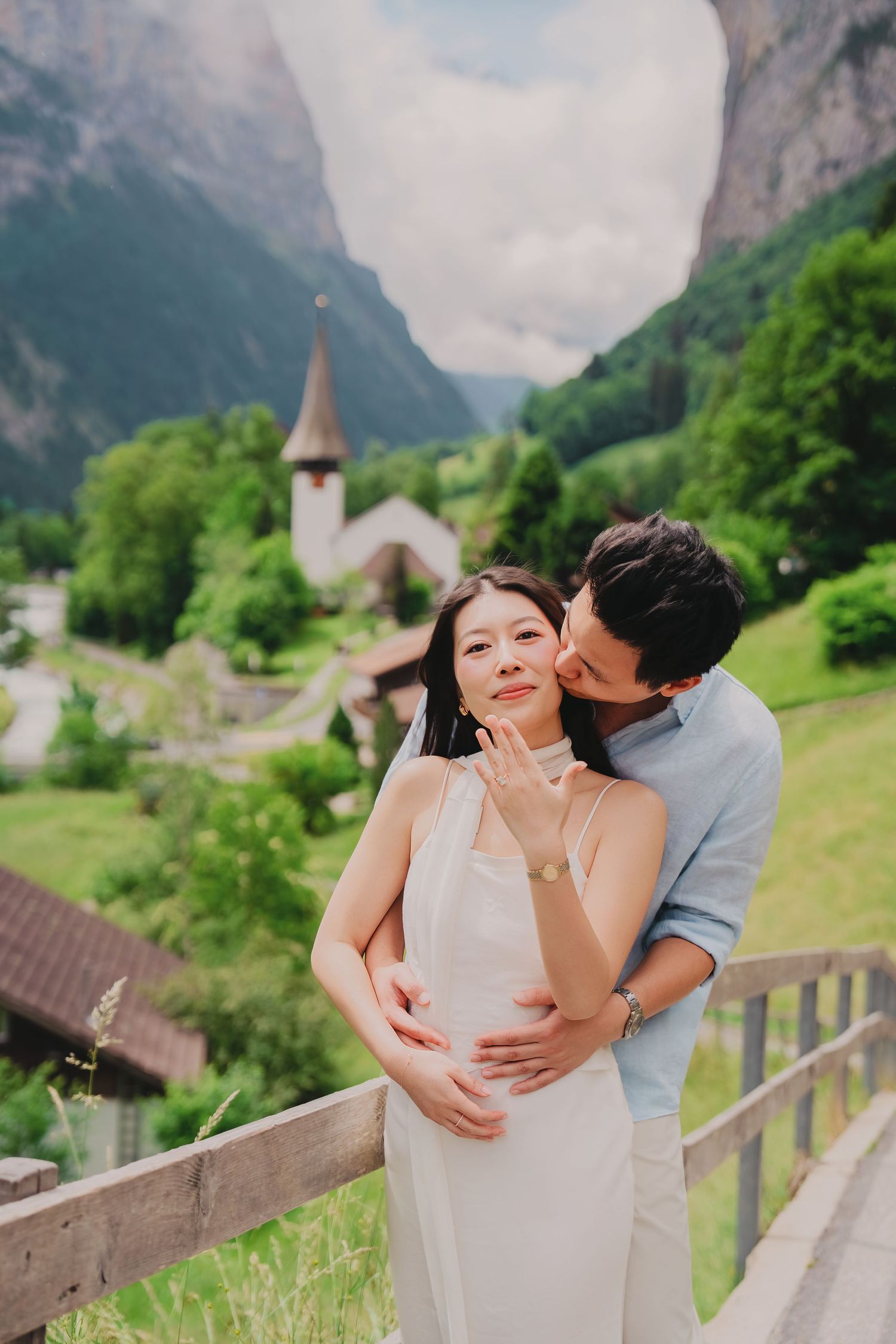 Man embracing woman from behind on a balcony overlooking a picturesque Swiss mountain village.