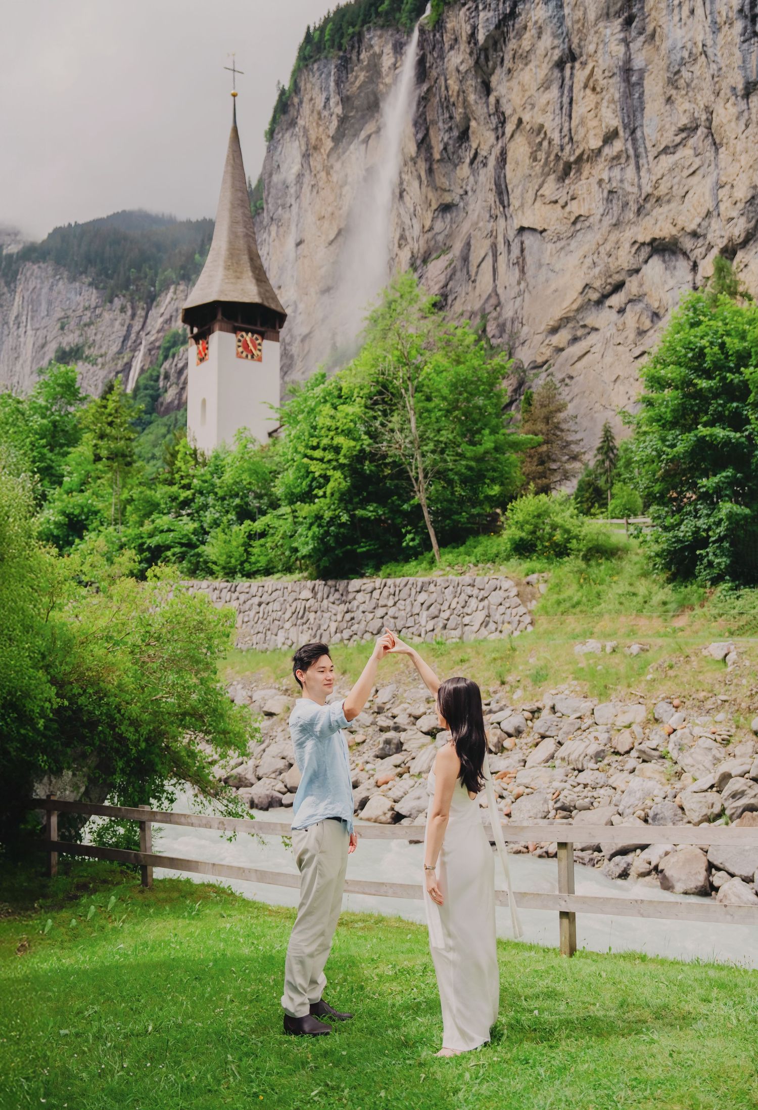 Couple dancing on green lawn with historic Swiss church tower and dramatic Lauterbrunnen waterfall behind them.