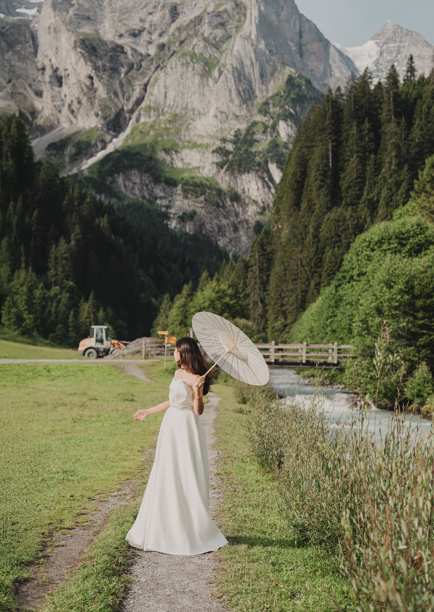 Bride in strapless white gown twirls parasol on scenic alpine path with majestic mountain views.