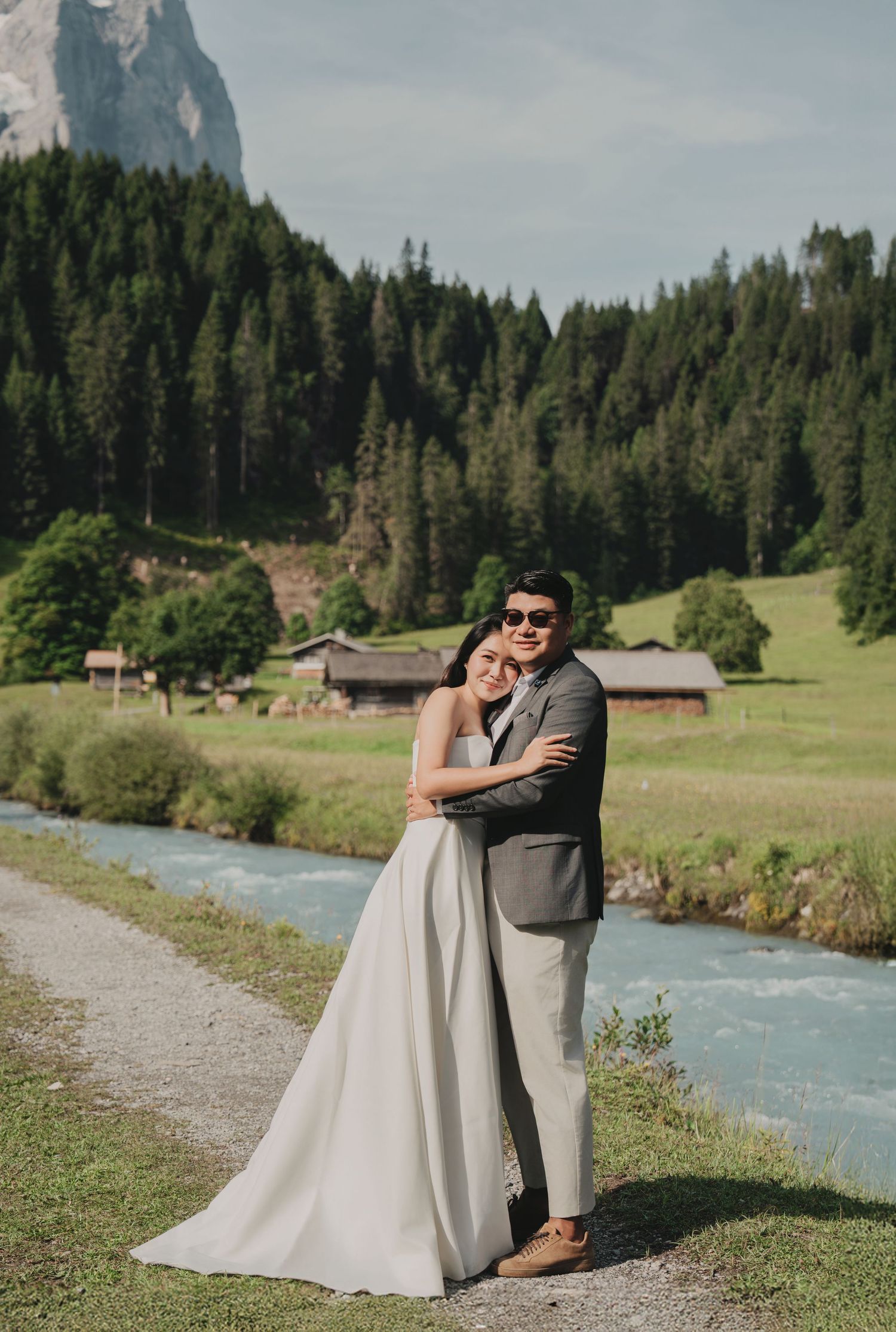 Couple in wedding attire embracing by a mountain stream in an Alpine valley with evergreen forests.