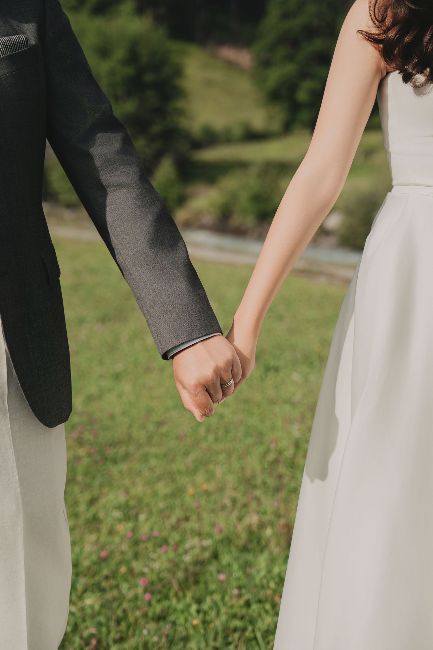 Close-up of bride and groom holding hands, bride in white gown and groom in gray suit outdoors.