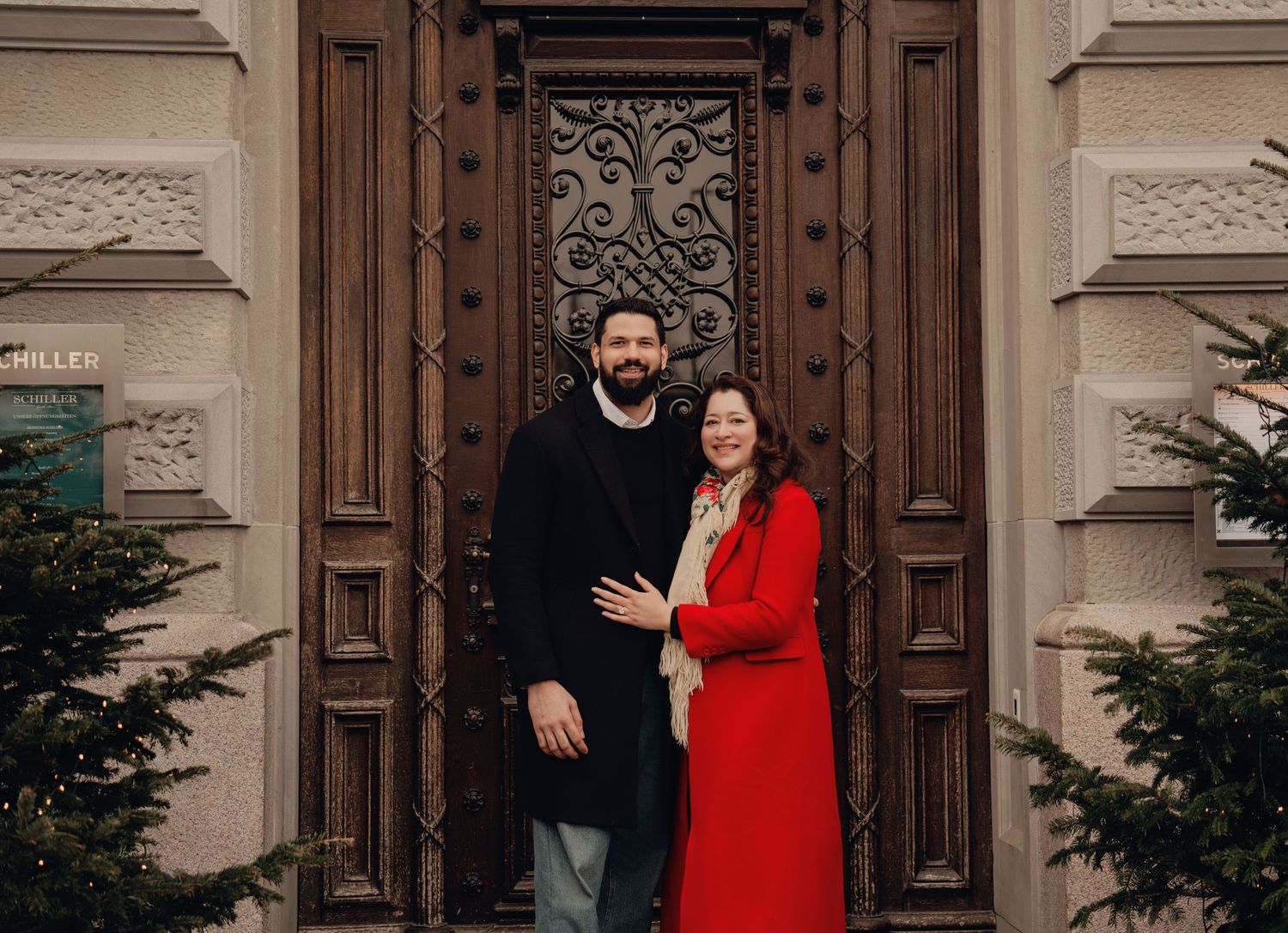 Smiling couple posing together in front of an elegant carved wooden door with Christmas trees.