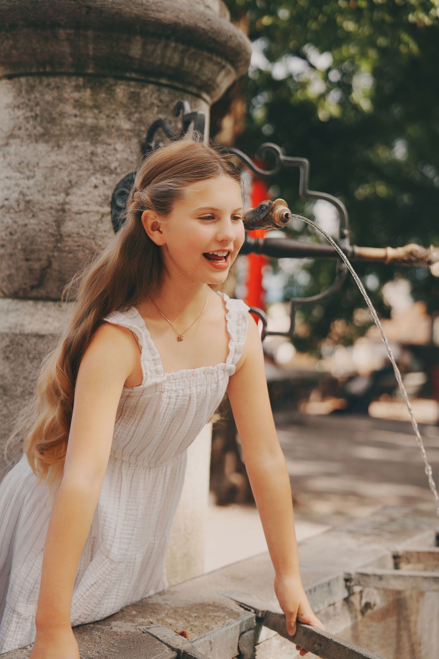 A laughing young girl in a white dress leans over an ornate stone fountain in a sunny European square.