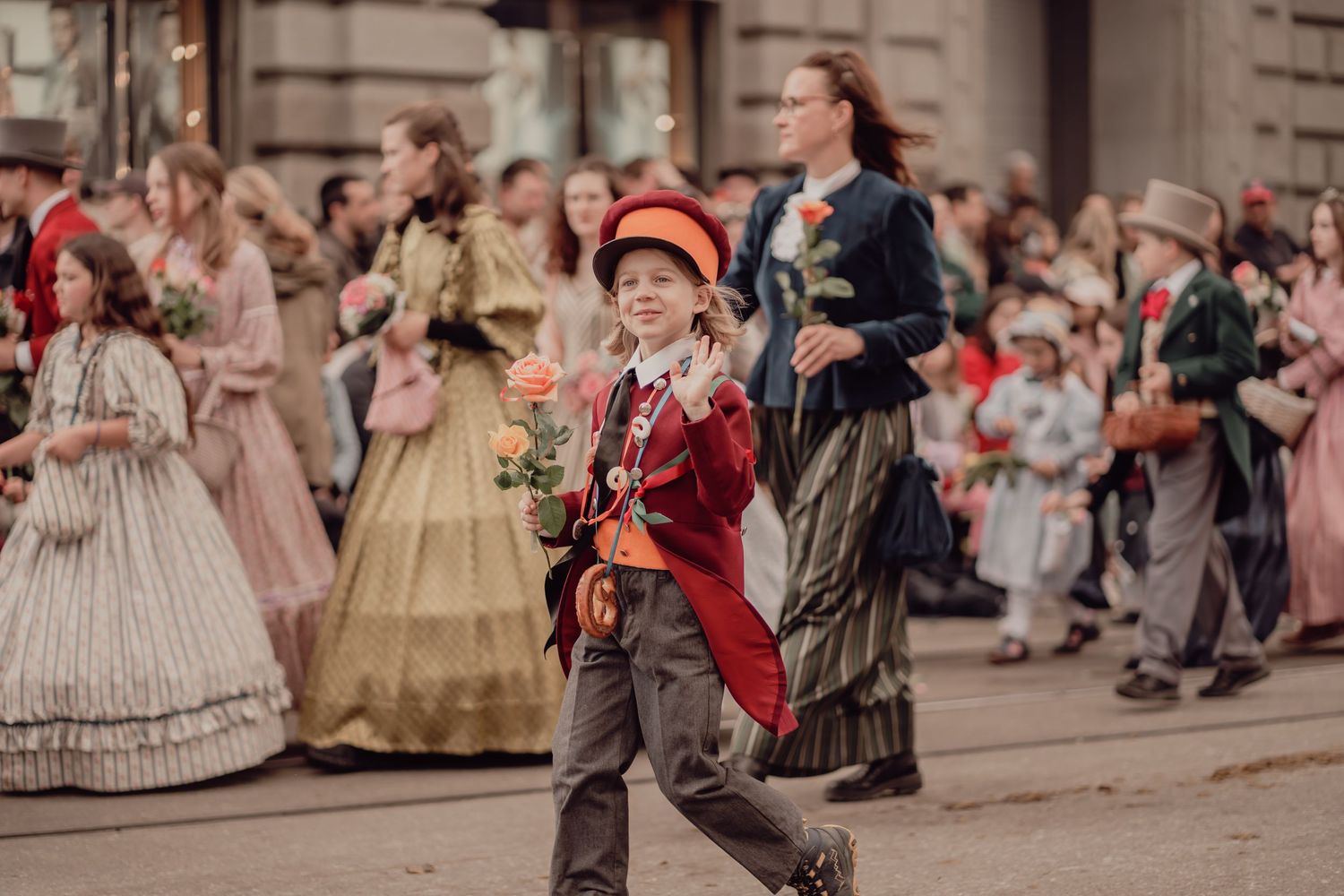 A young boy in a red Victorian suit carries a rose while marching in a historical costume parade.