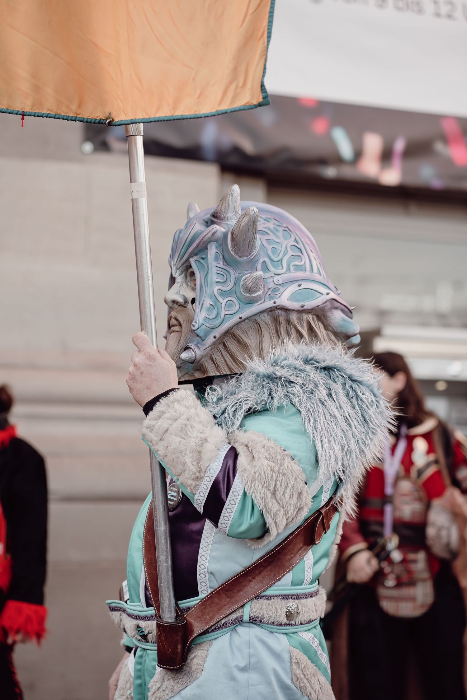 Person in teal dragon warrior costume carries a flag at a colorful carnival parade.