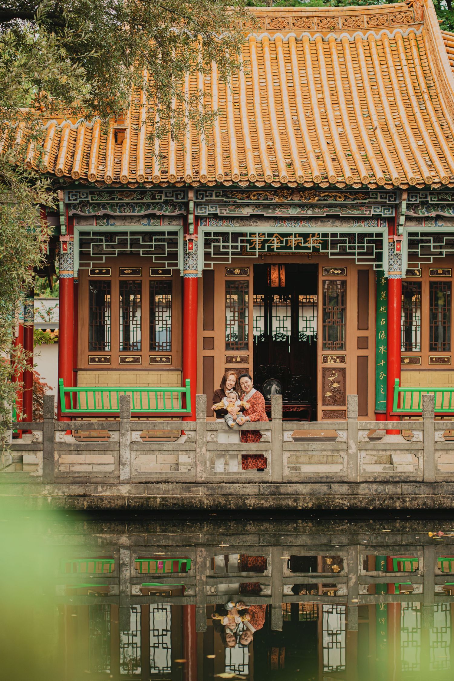 Couple holding baby on ornate Chinese temple veranda reflected beautifully in calm green pond.