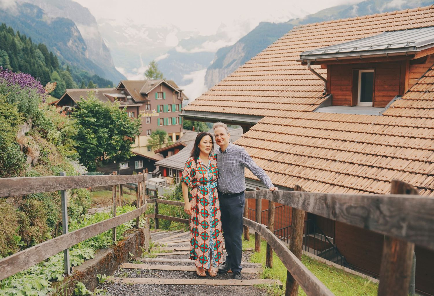 Couple embraces on wooden steps with Swiss chalet architecture and misty mountain peaks behind them.