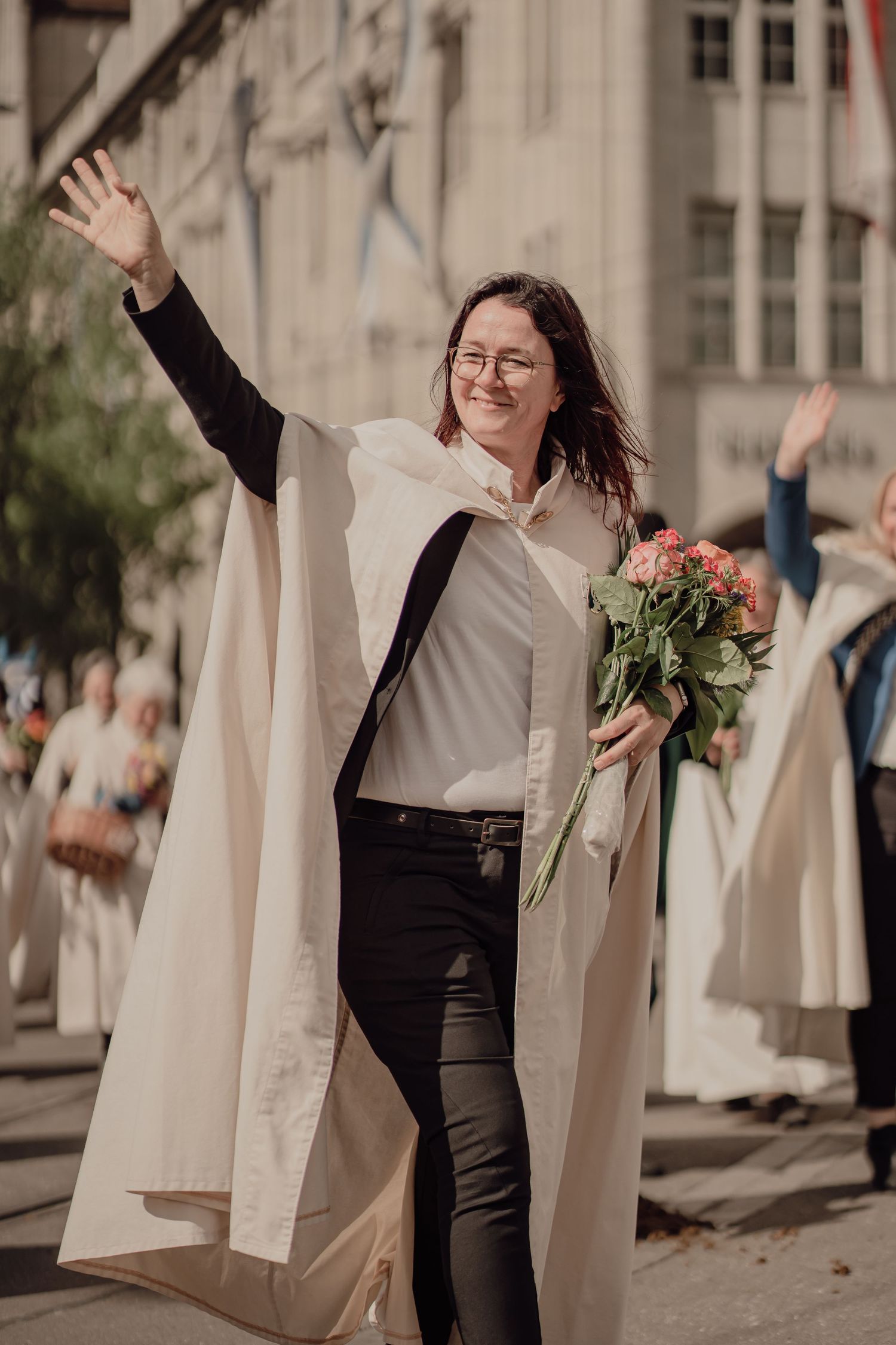 Smiling woman in white ceremonial cape waves to crowd while holding pink bouquet at street parade.