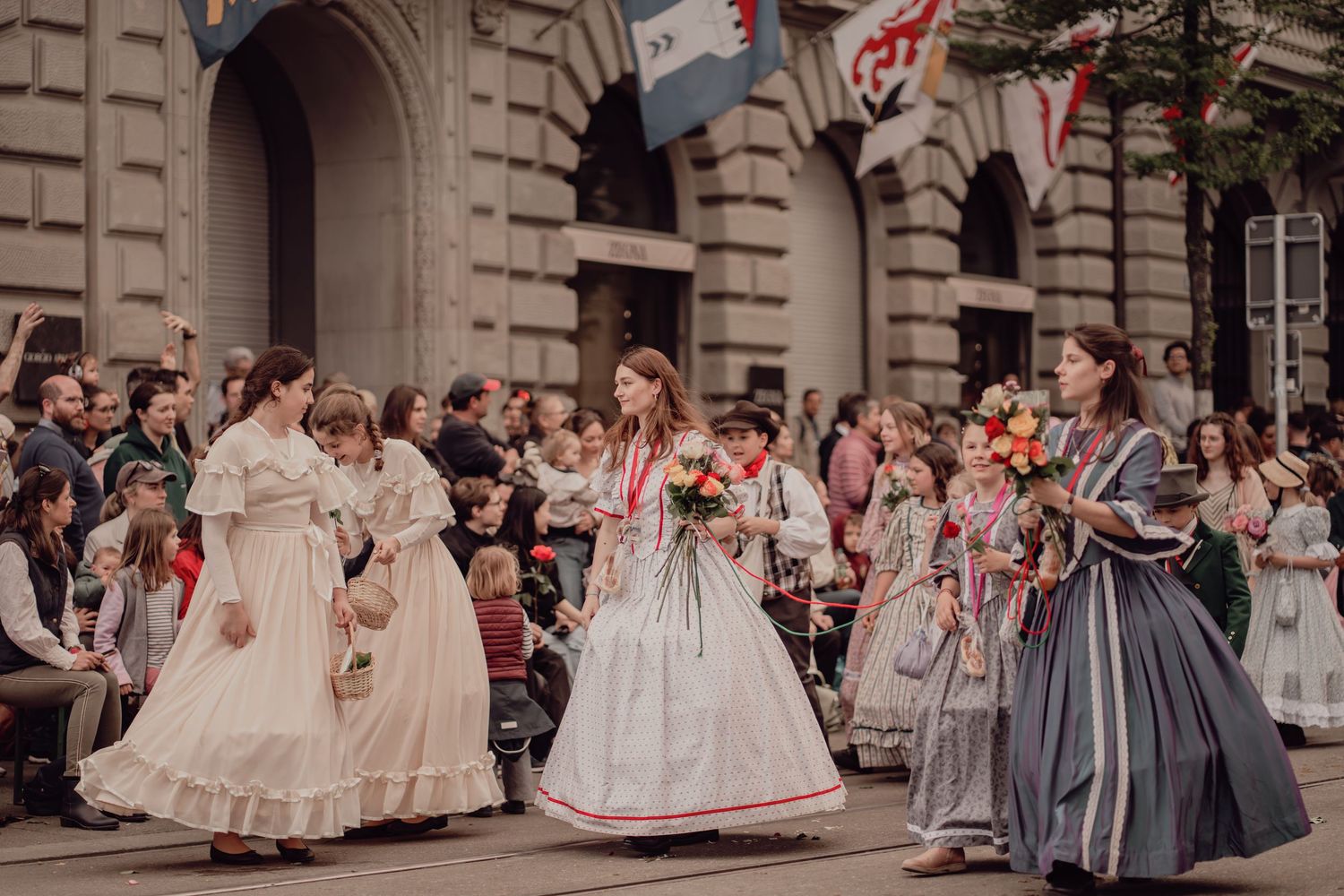 Girls and women in Victorian-era costumes carry flower bouquets during an elegant historical street parade.