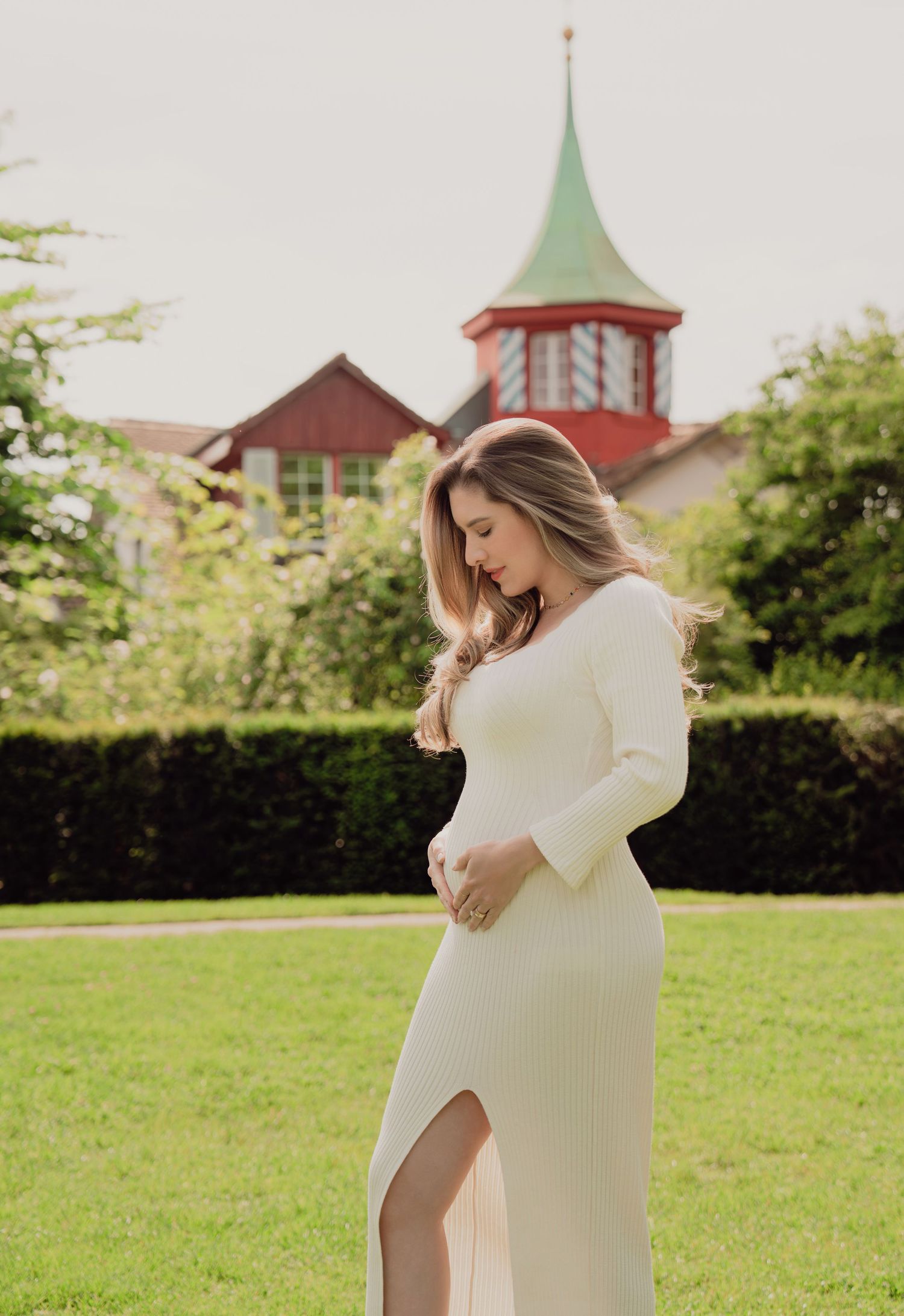Pregnant woman in a cream dress cradling her baby bump in a garden with a red building backdrop.