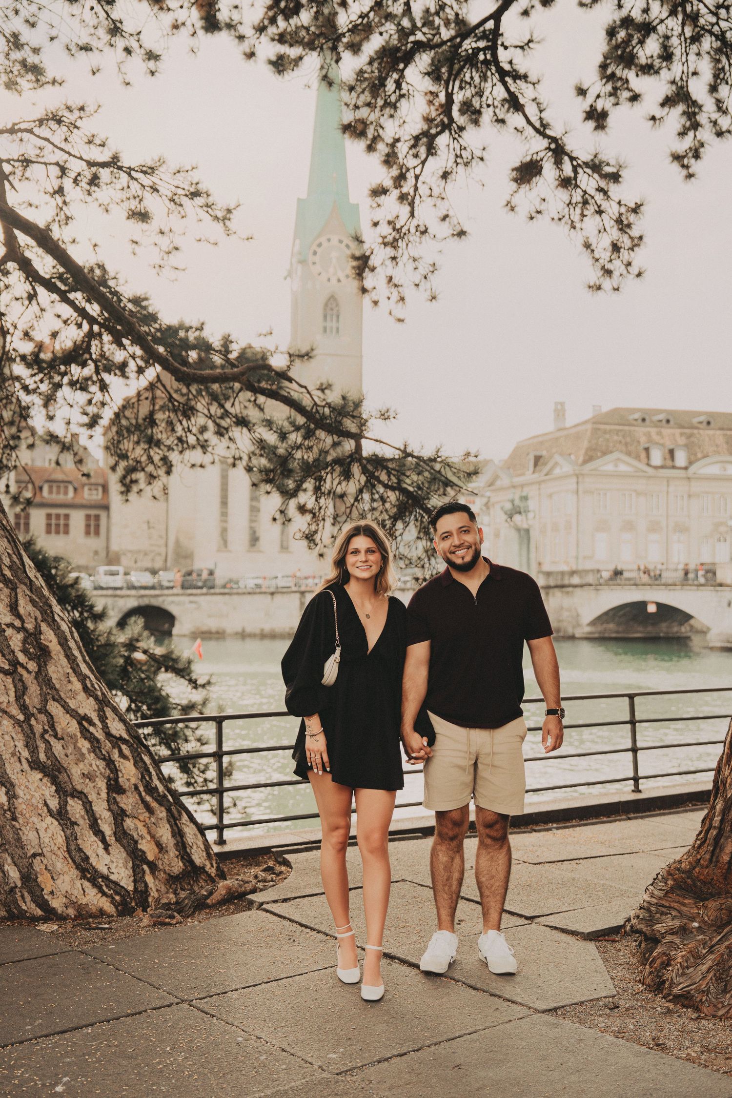 Romantic couple holding hands under tree branches by Zurich riverfront with church spire behind.