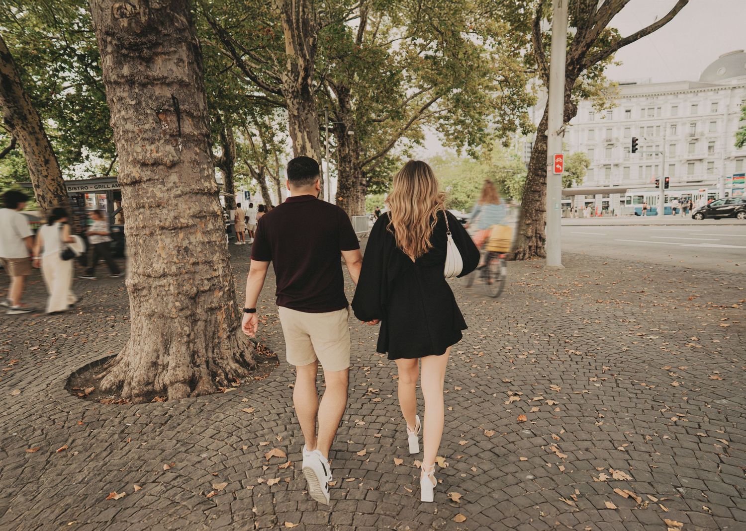 Couple walking hand in hand through a tree-lined cobblestone plaza in a European city.