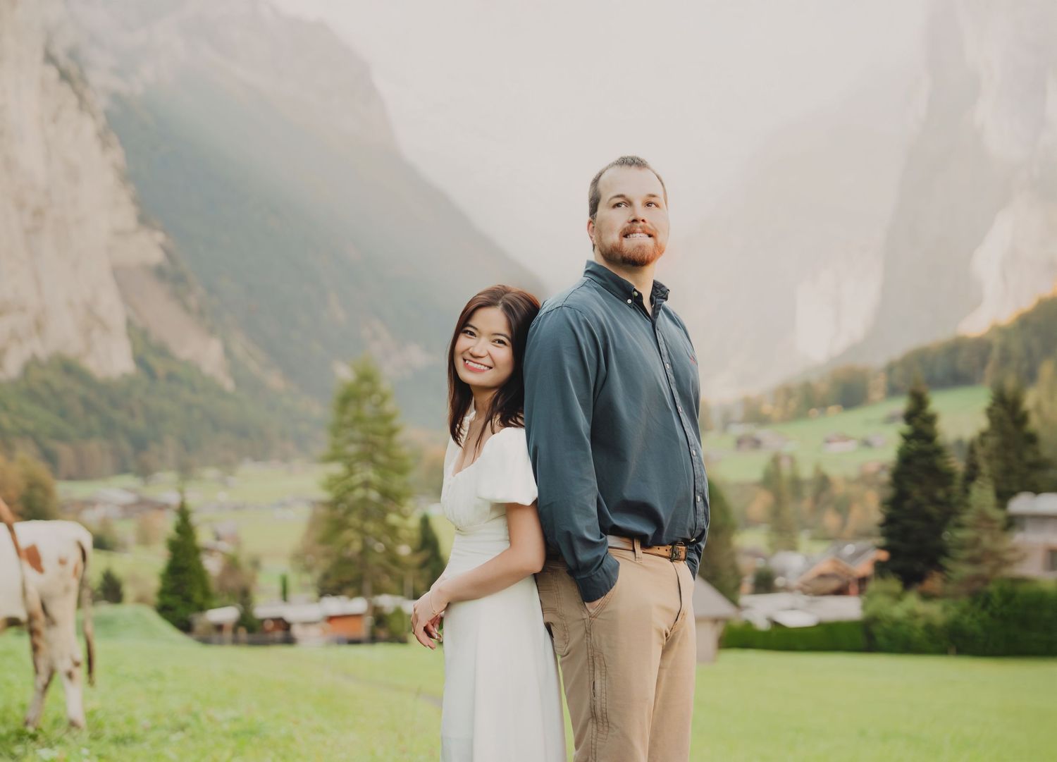 Couple posing back-to-back in a lush Swiss valley with dramatic mountain cliffs and alpine village in the background.
