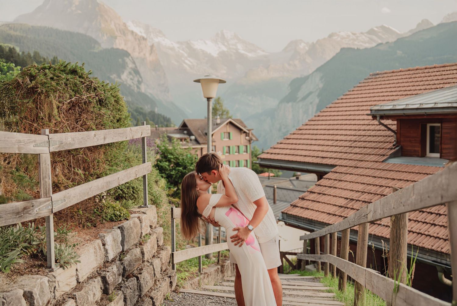Couple kissing on stone steps in Swiss alpine village with snow-capped mountains backdrop.