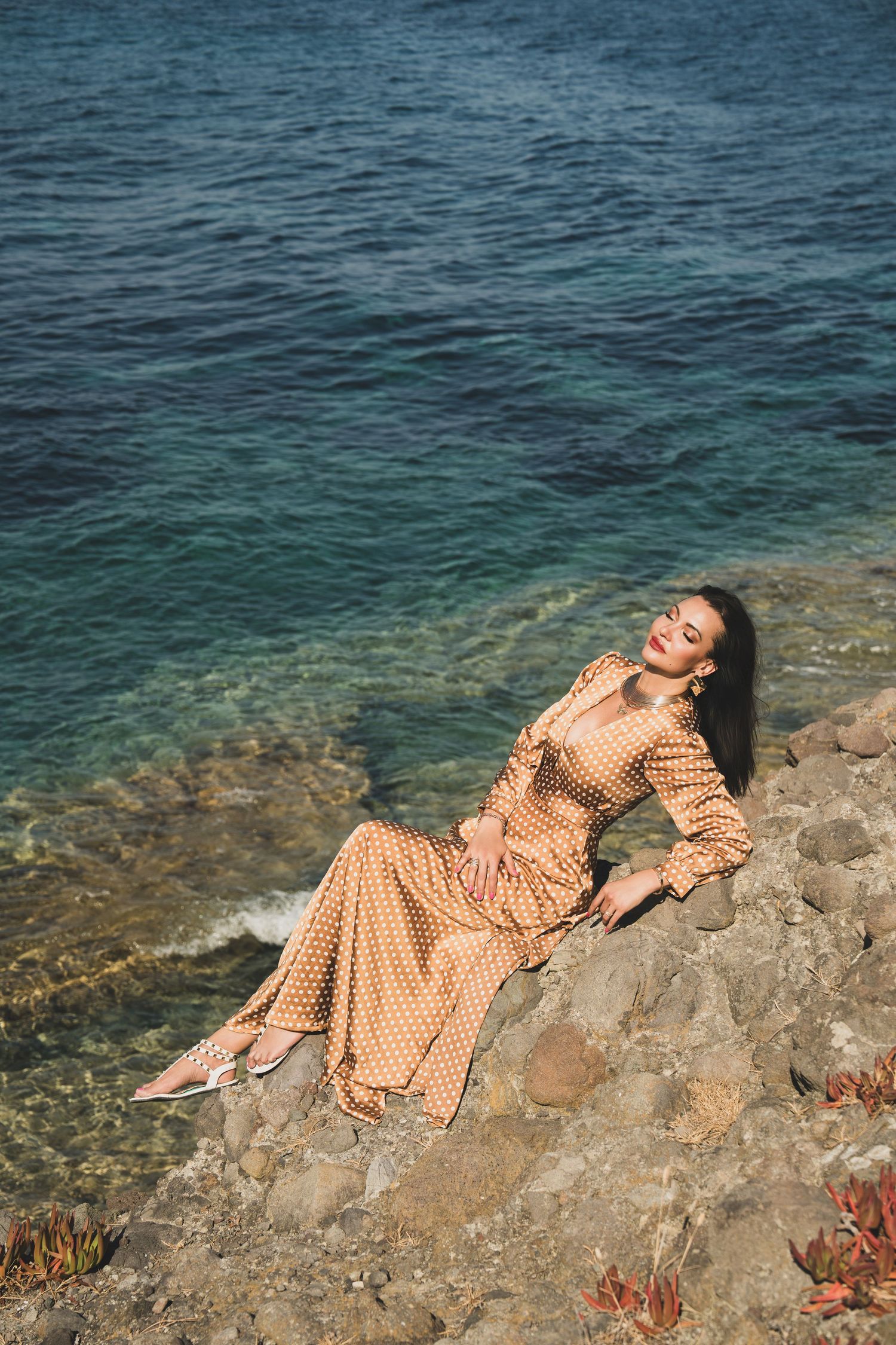 Woman in tan polka dot maxi dress posing on rocky seaside cliff with turquoise water backdrop.