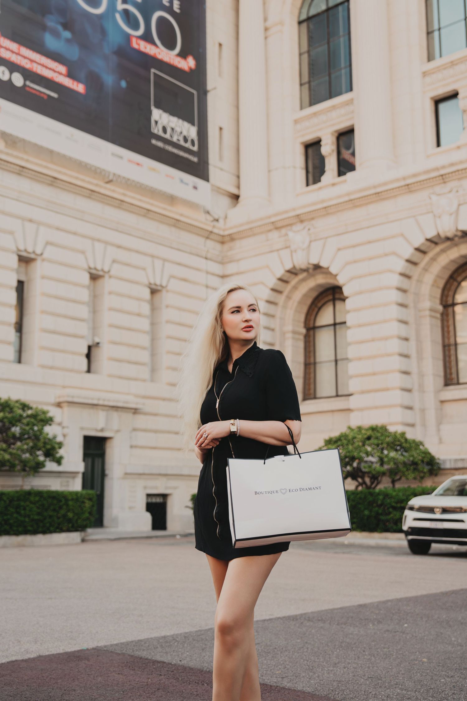 Blonde woman in black dress holding Bottega Veneta shopping bag outside elegant building.