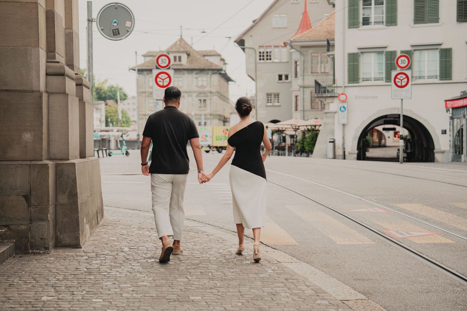 Couple walking hand in hand through Zurich's historic cobblestone streets past tram signs.