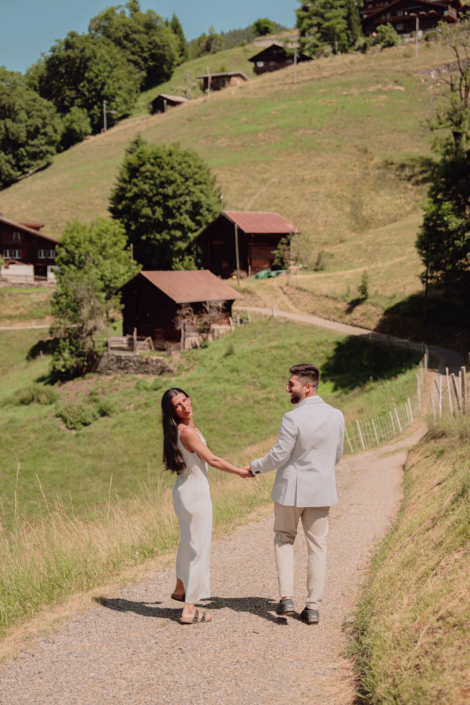 Bride in white dress and groom in light suit holding hands on a gravel path with Alpine chalets.