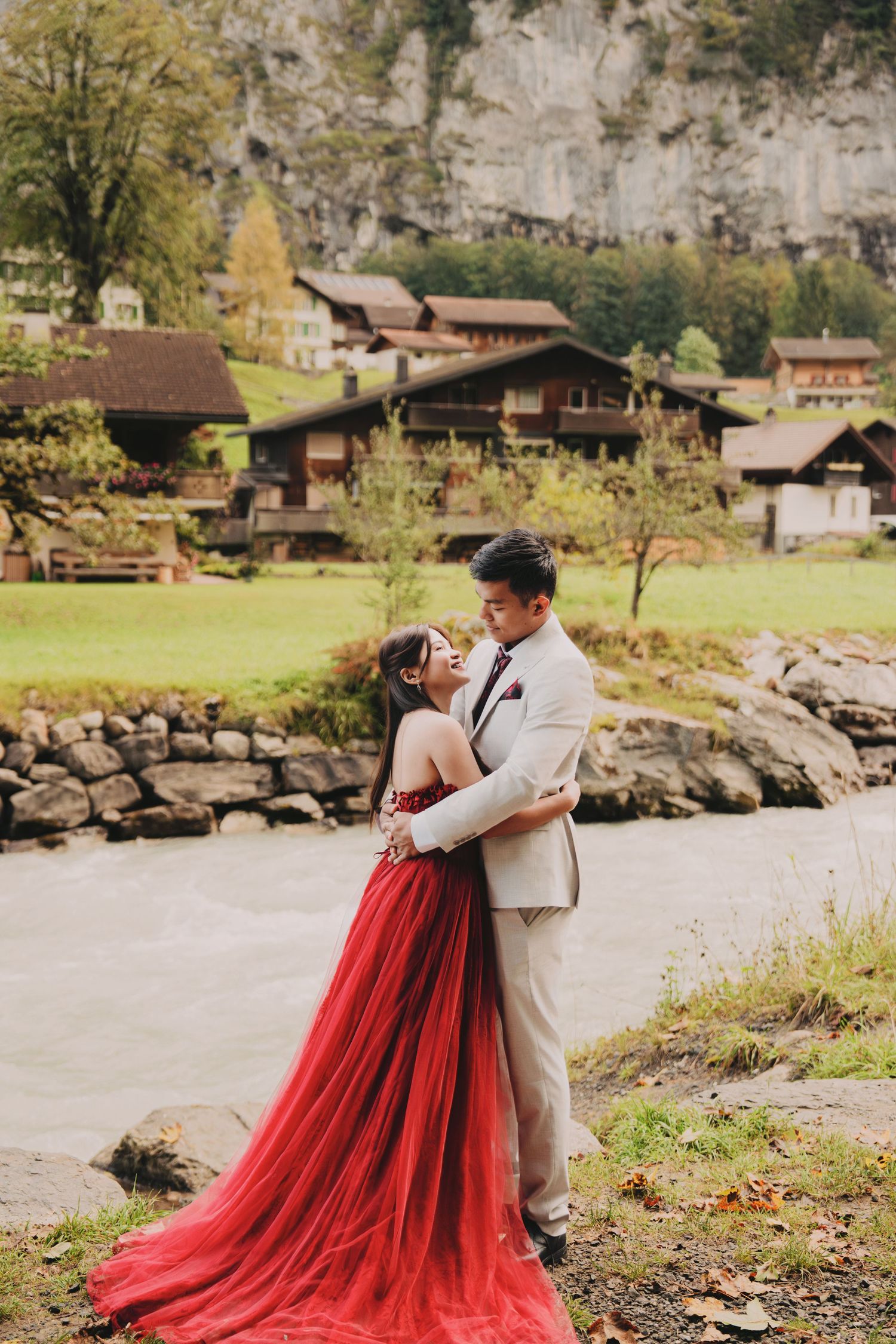 Couple embracing by a river in Lauterbrunnen, Switzerland, bride in red gown and groom in beige suit.