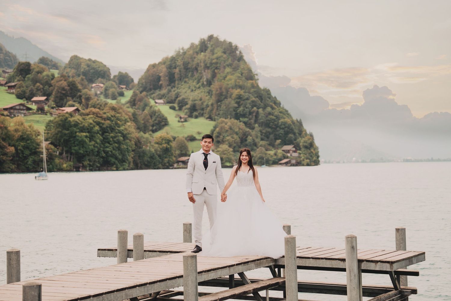 Newlyweds holding hands walking on wooden dock with green hillside and alpine lake in background.