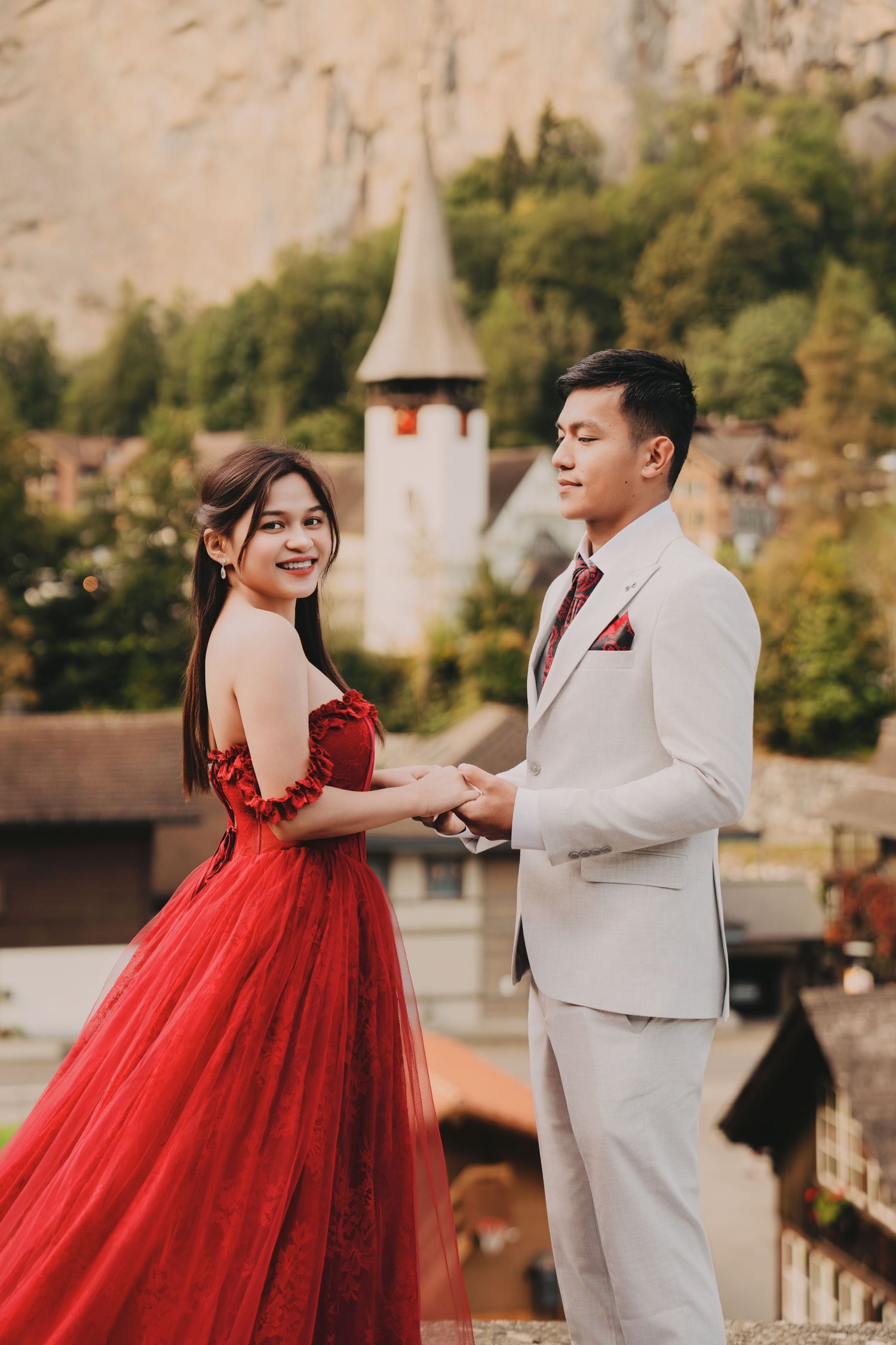 Smiling bride in red tulle gown holds groom's hands near alpine church in Switzerland.