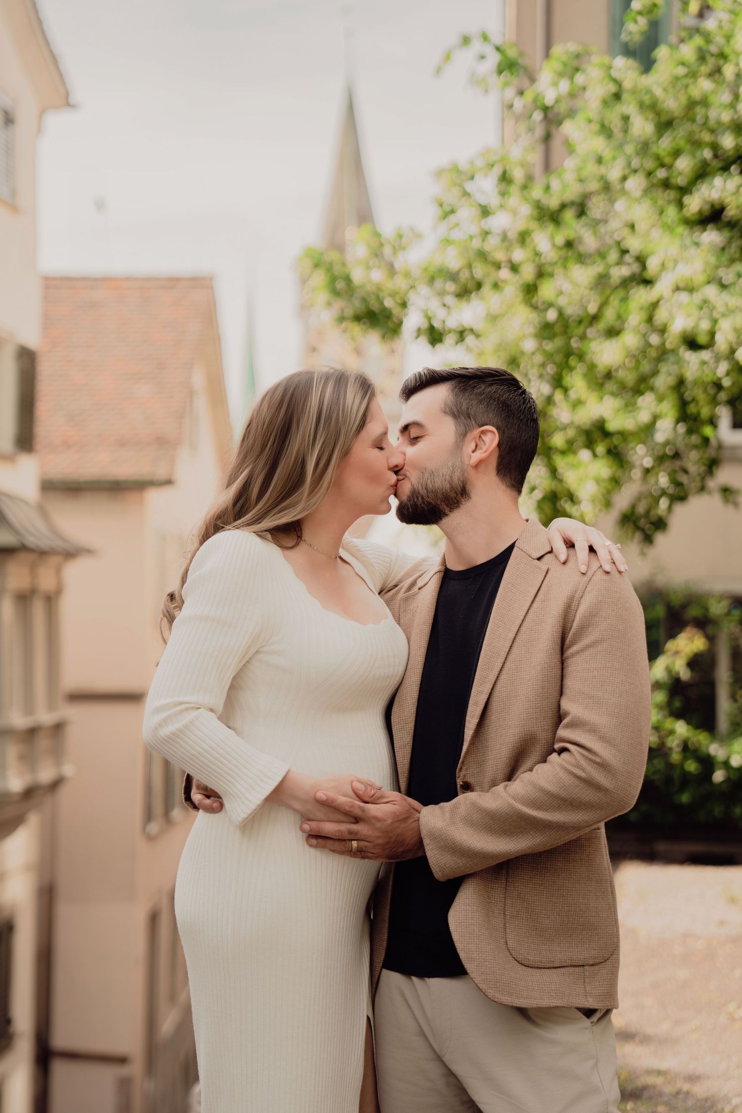 Expecting couple shares a romantic kiss while man tenderly holds pregnant belly in Zurich old town setting.
