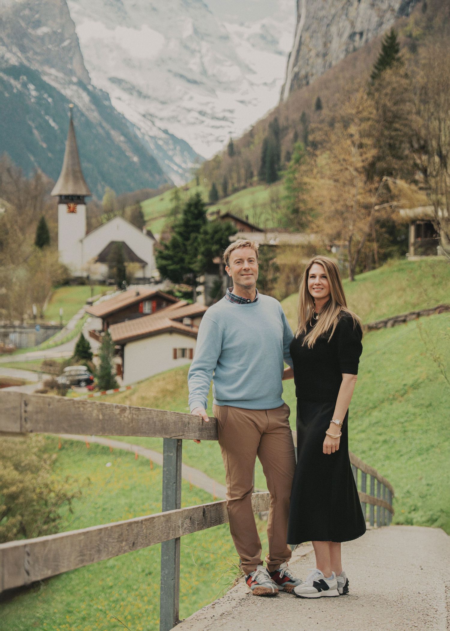 Couple posing together on a wooden fence with a Swiss mountain village church and snow-capped Alps behind them.