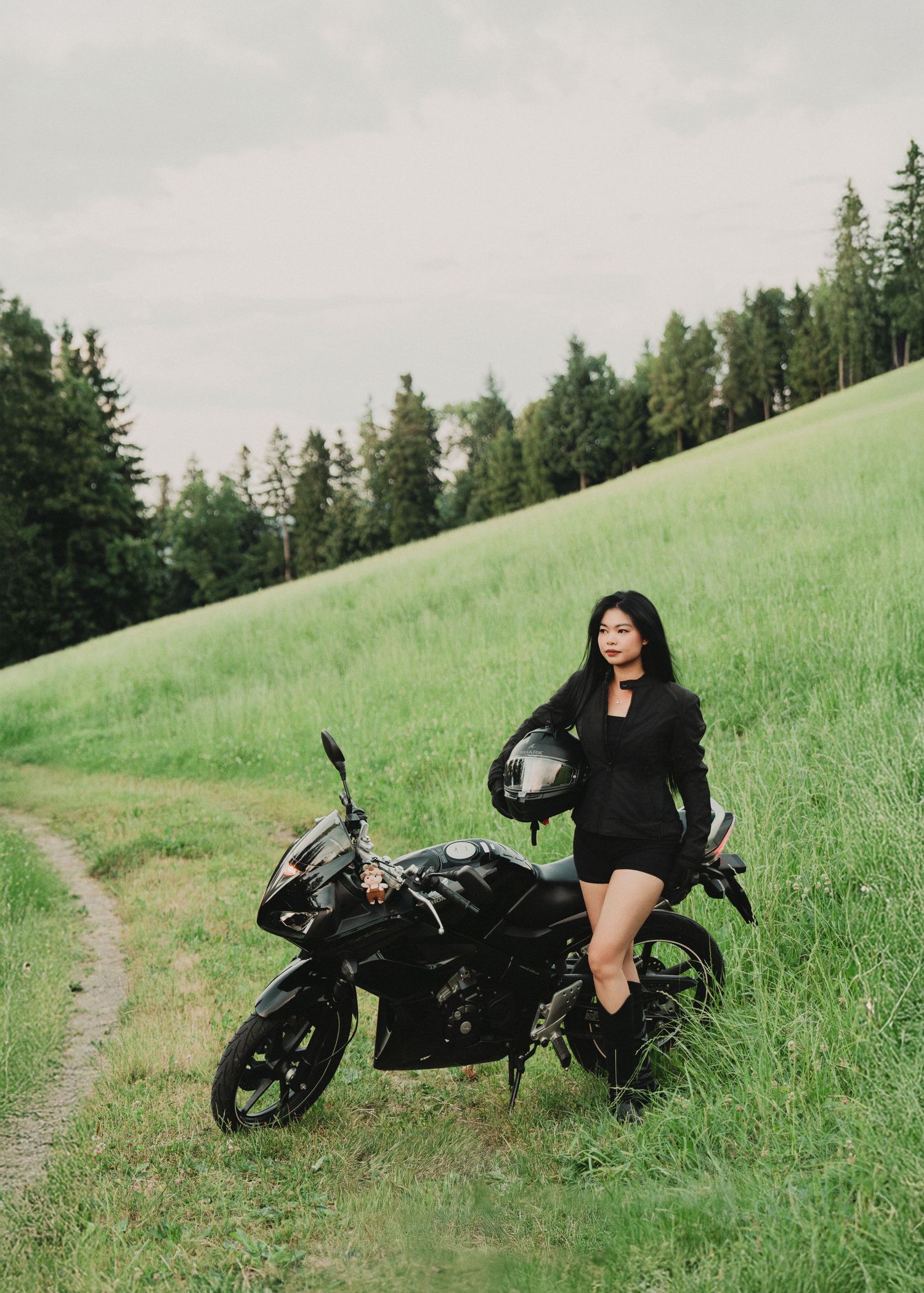 Female rider in black shorts and jacket leaning on black motorcycle on scenic green hillside.