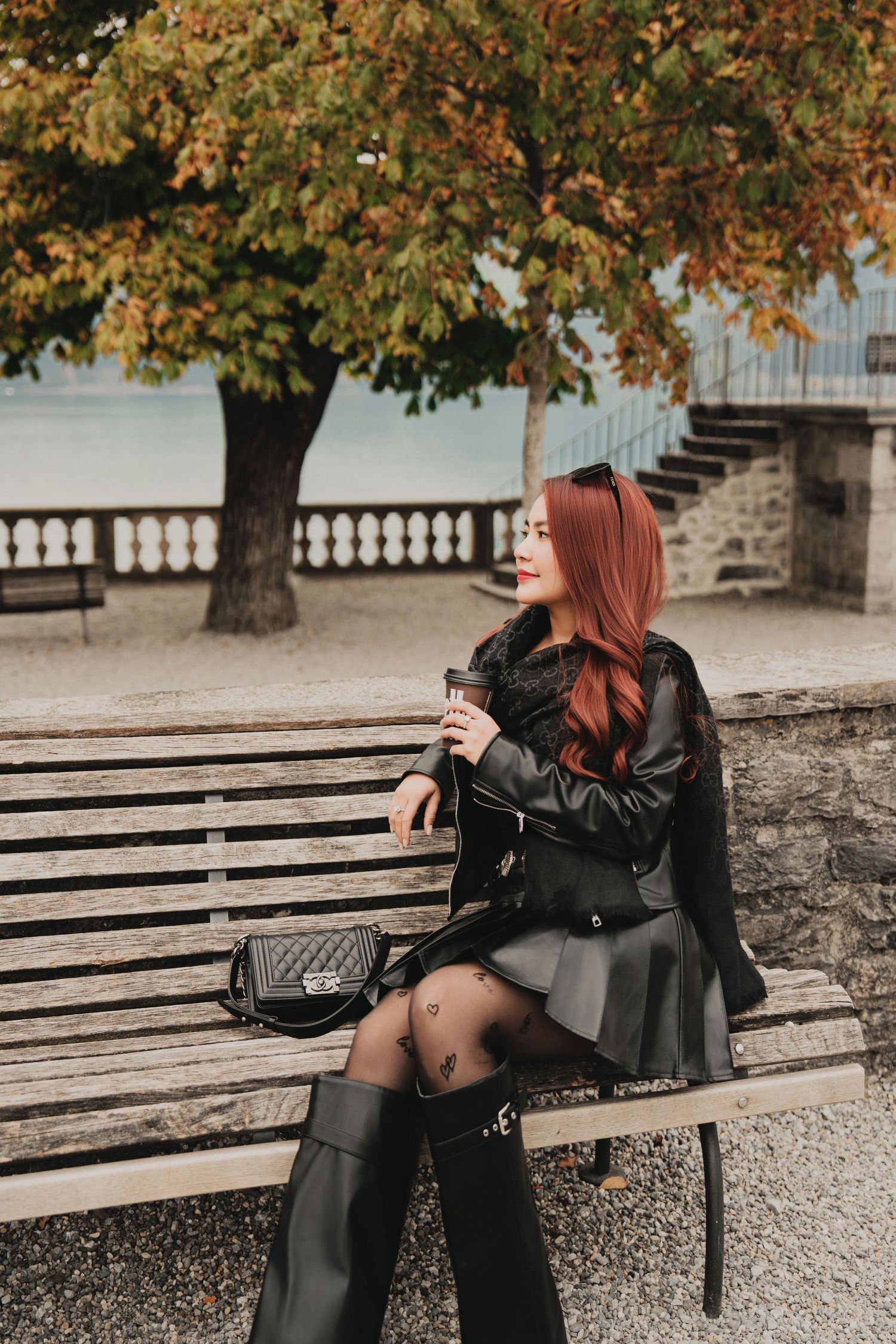 Red-haired woman in black leather jacket and skirt sitting on a bench in an autumn park.