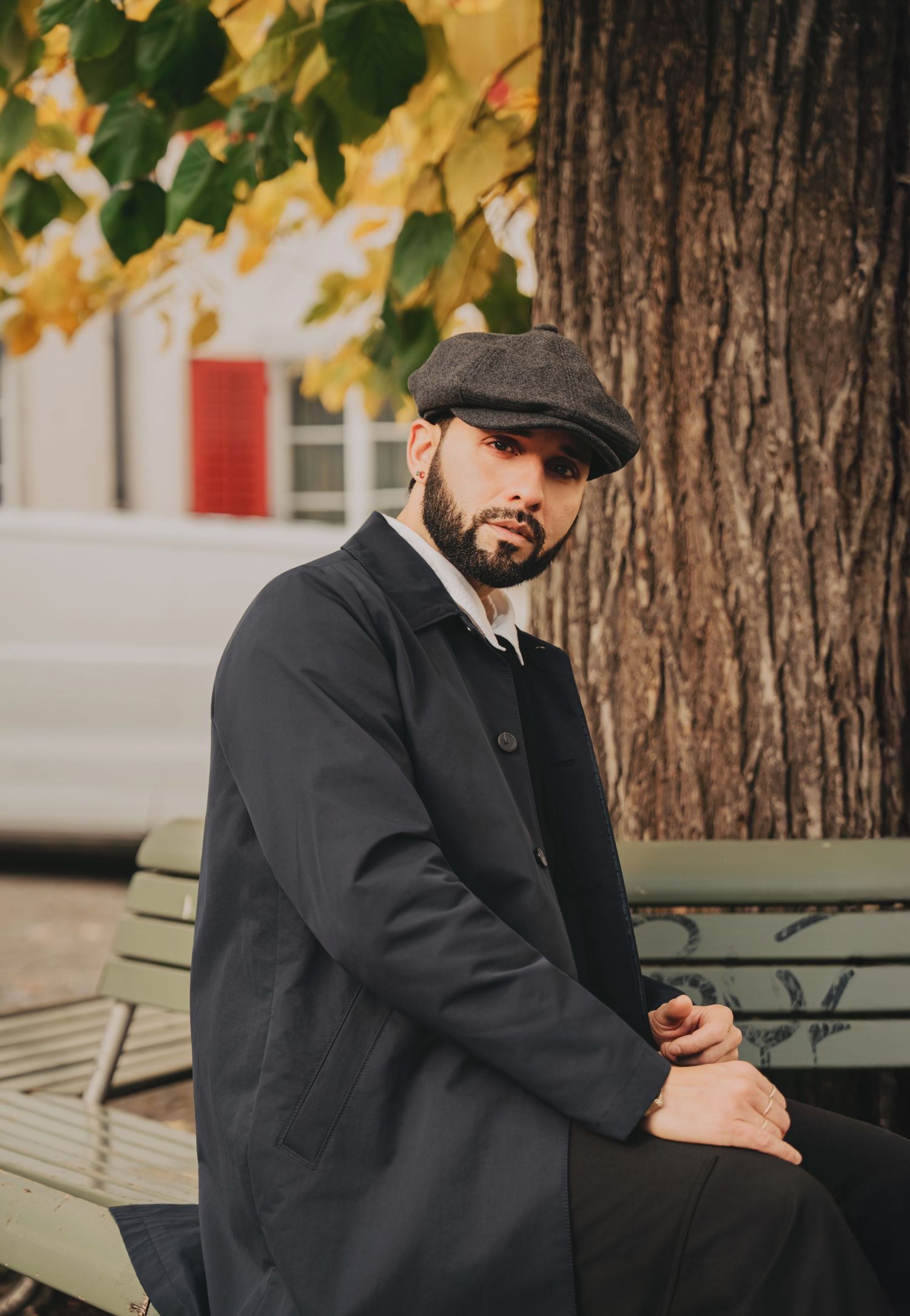 Stylish bearded man in dark coat and flat cap sitting on bench under golden autumn tree looking at camera.