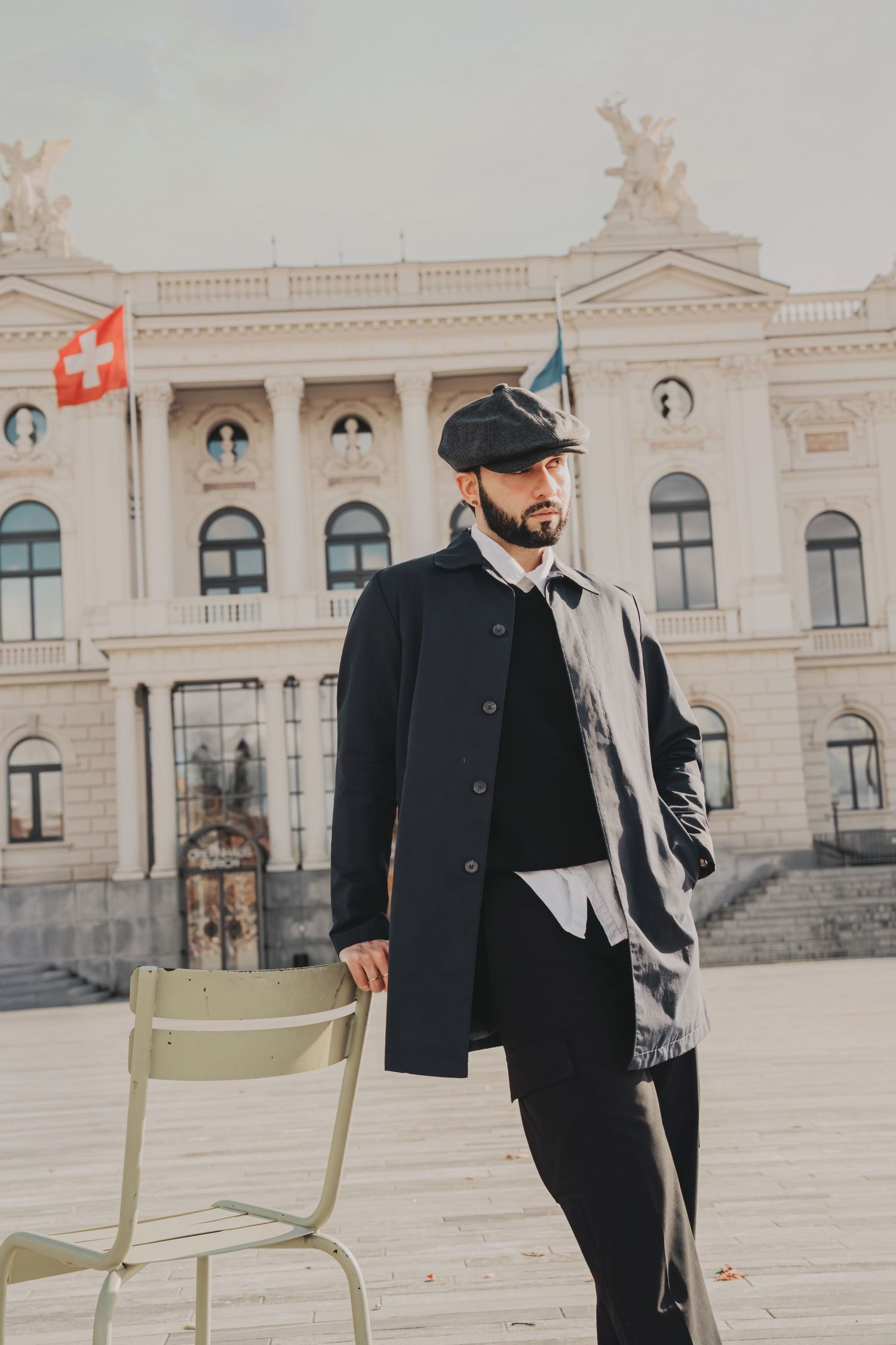 Dapper bearded man in black coat and flat cap standing before ornate European building with Swiss flag.