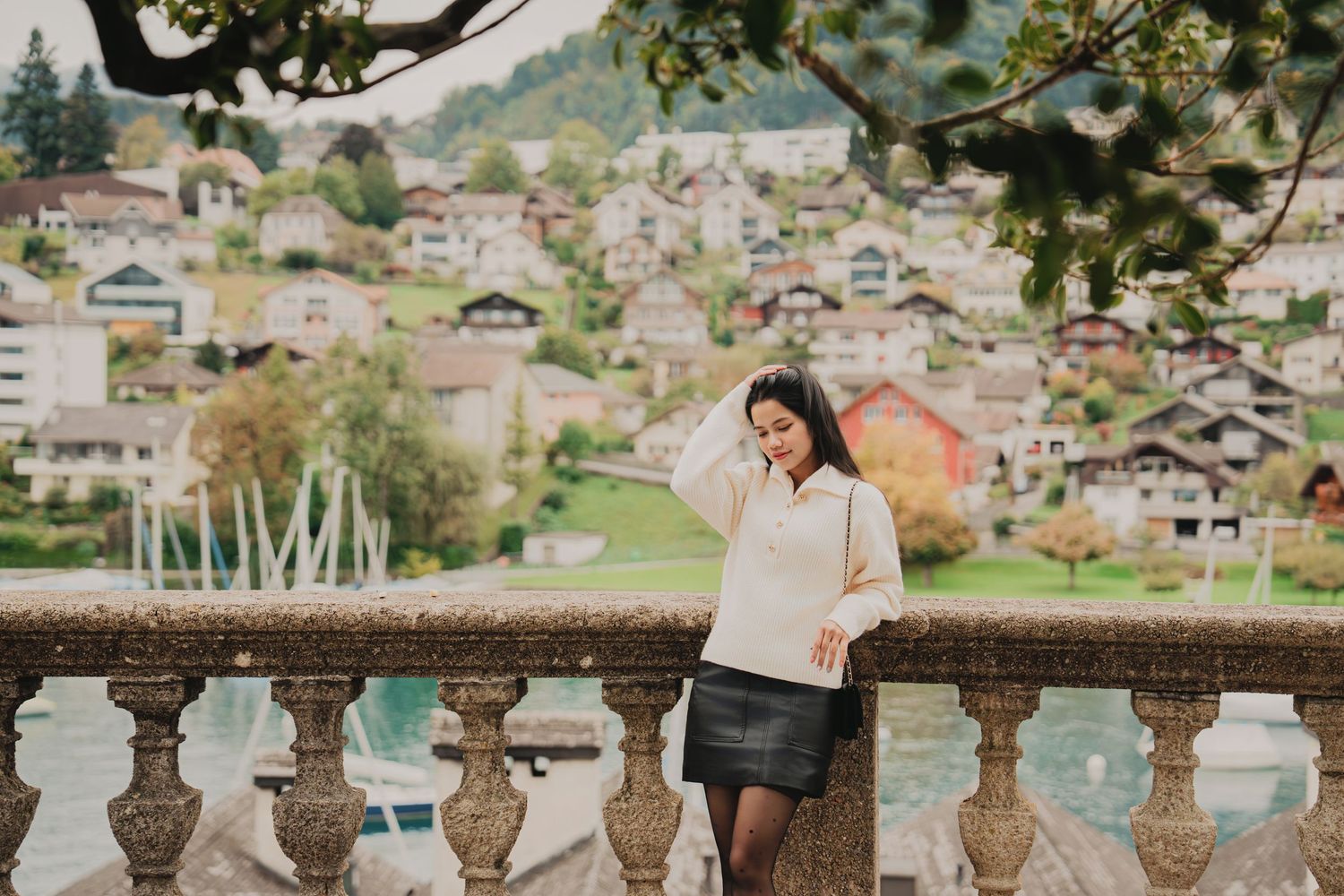 Smiling woman in cream sweater and black skirt poses by ornate stone balustrade with Swiss village view.