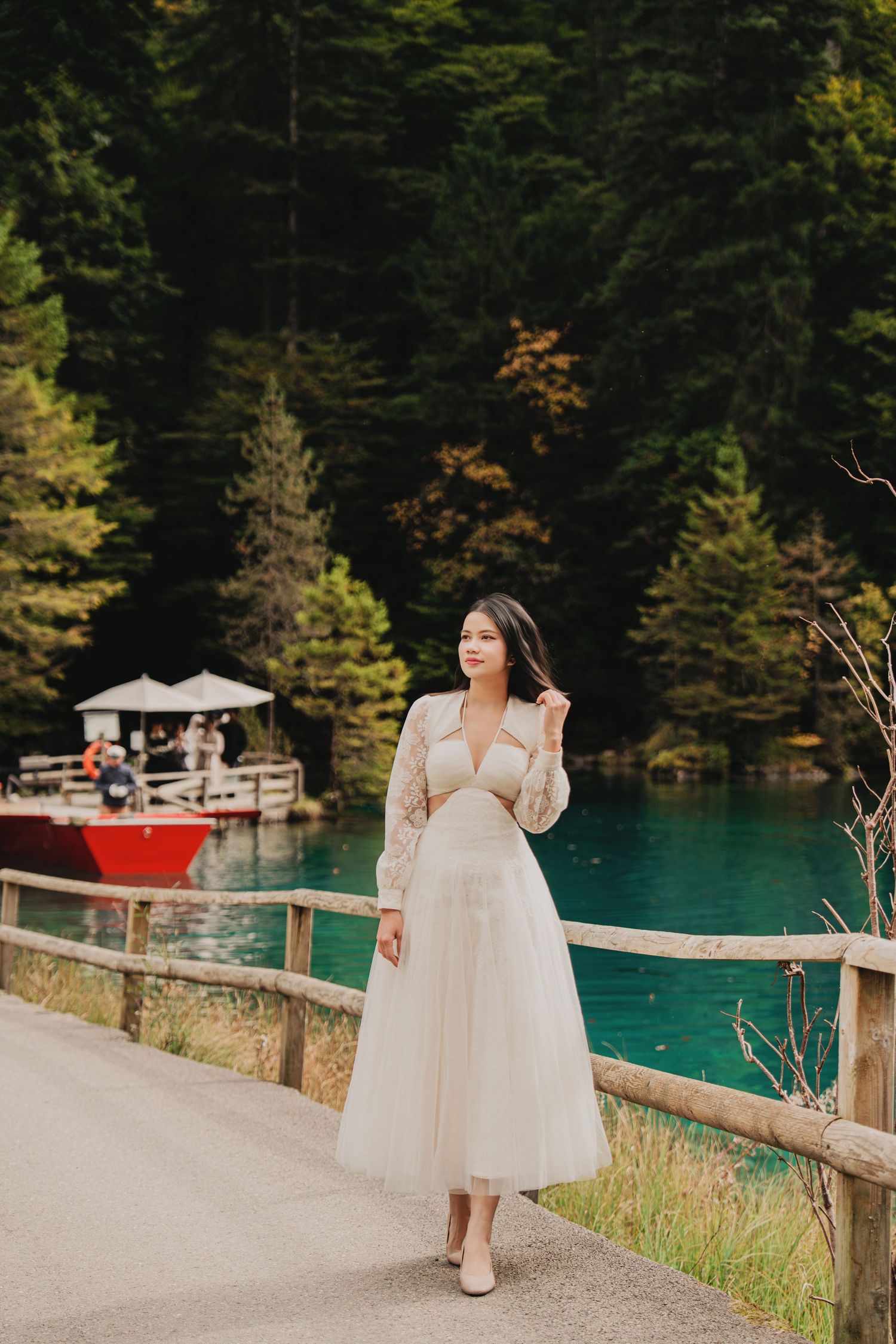 Woman in white tulle midi dress posing by wooden fence with turquoise lake and red boat in background.