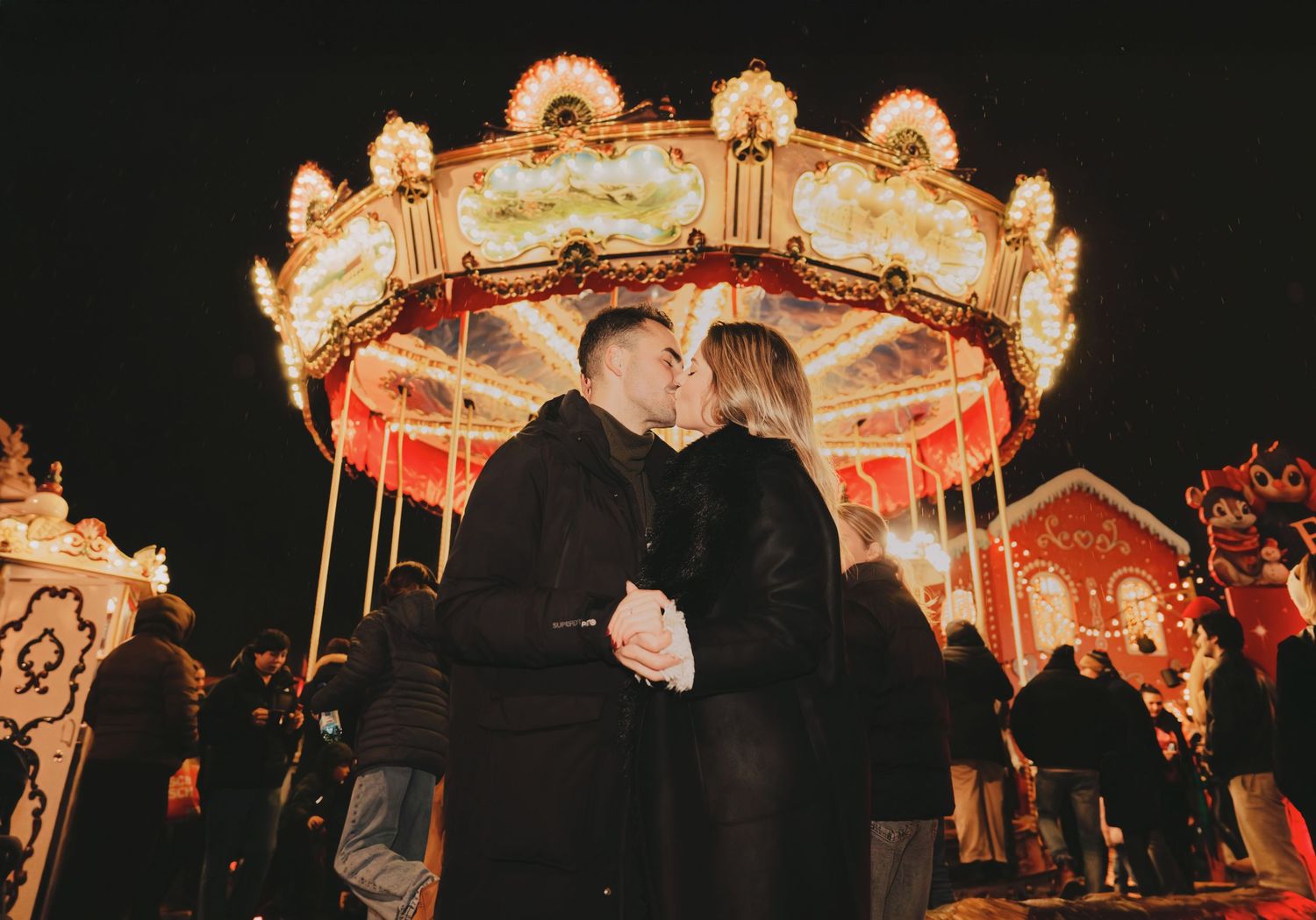 Couple kissing in front of a beautifully lit carousel at a festive Christmas market at night.