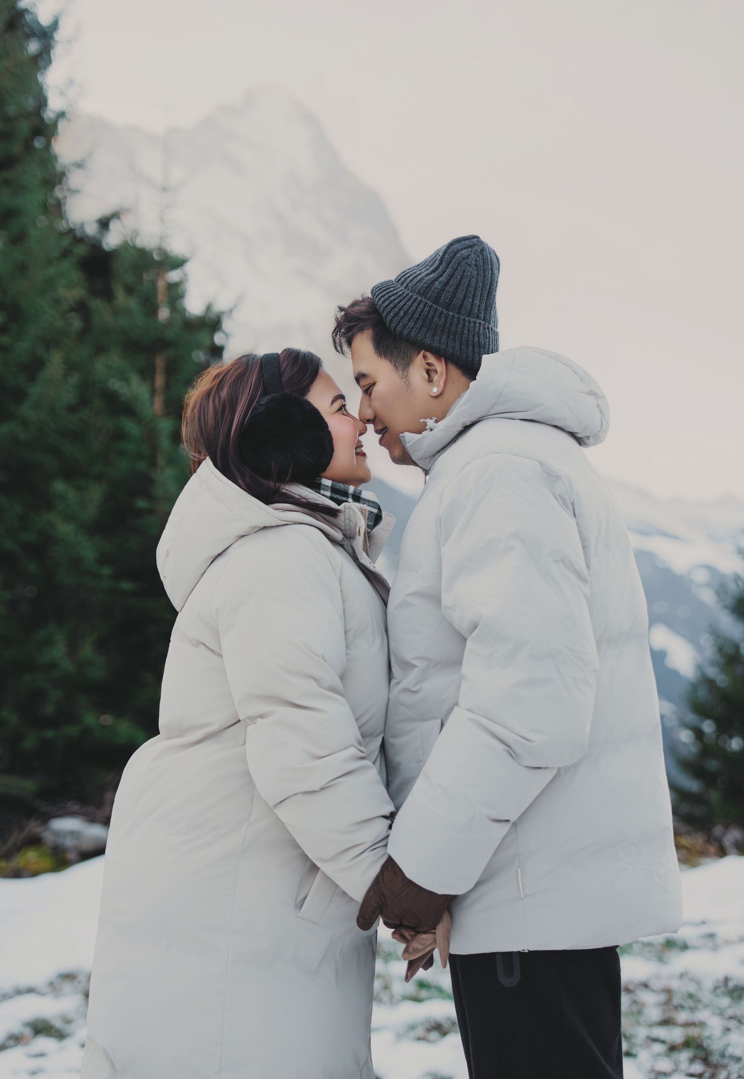 Couple in white puffer jackets sharing a romantic moment with snowy mountain forest backdrop.