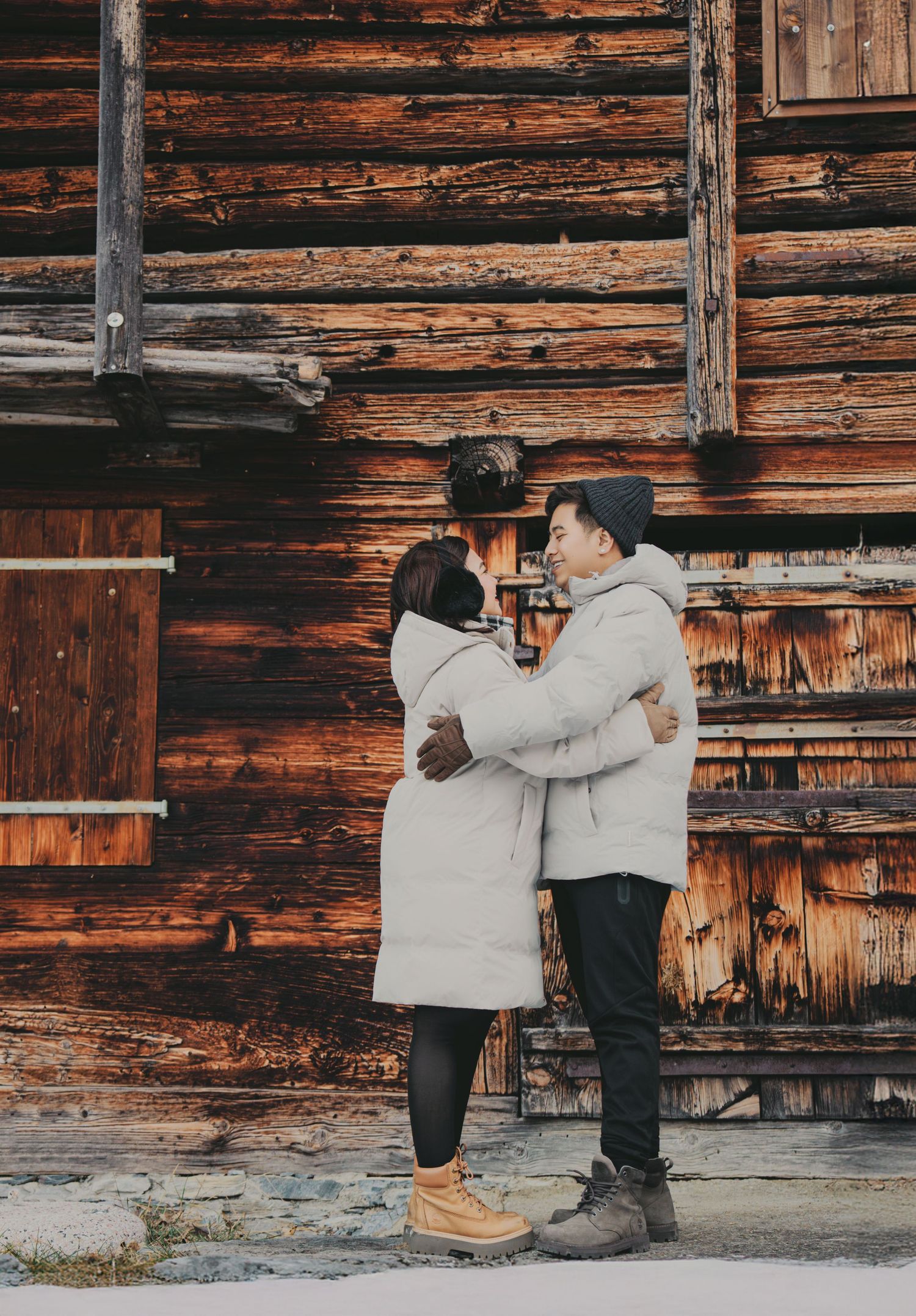 Couple embracing in white puffer coats before rustic wooden log cabin wall in winter.