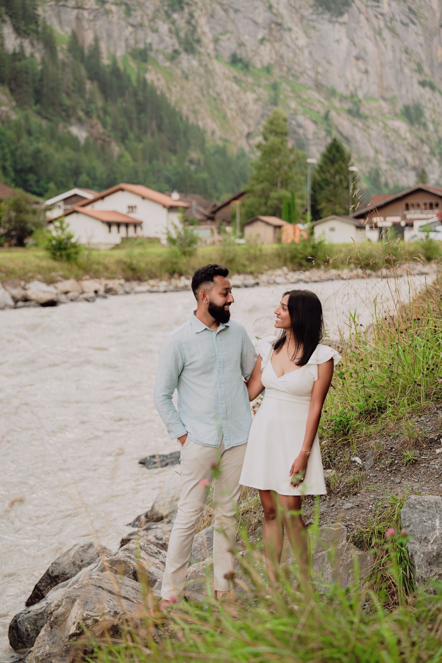 Couple standing together on rocky riverbank with Swiss Alpine village and towering mountain cliffs behind them.