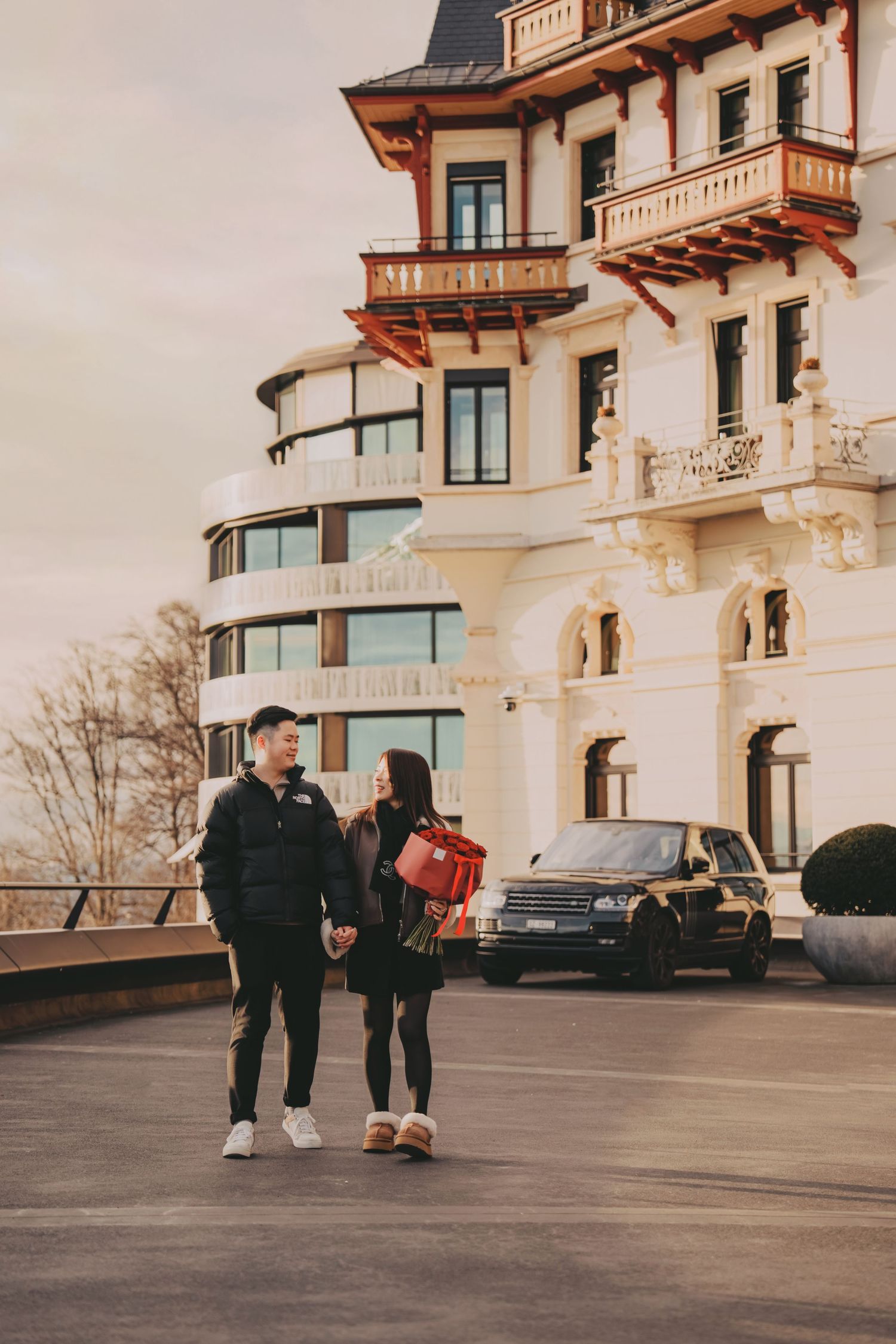 Couple walking hand in hand outside a grand European hotel with a black Range Rover parked nearby.