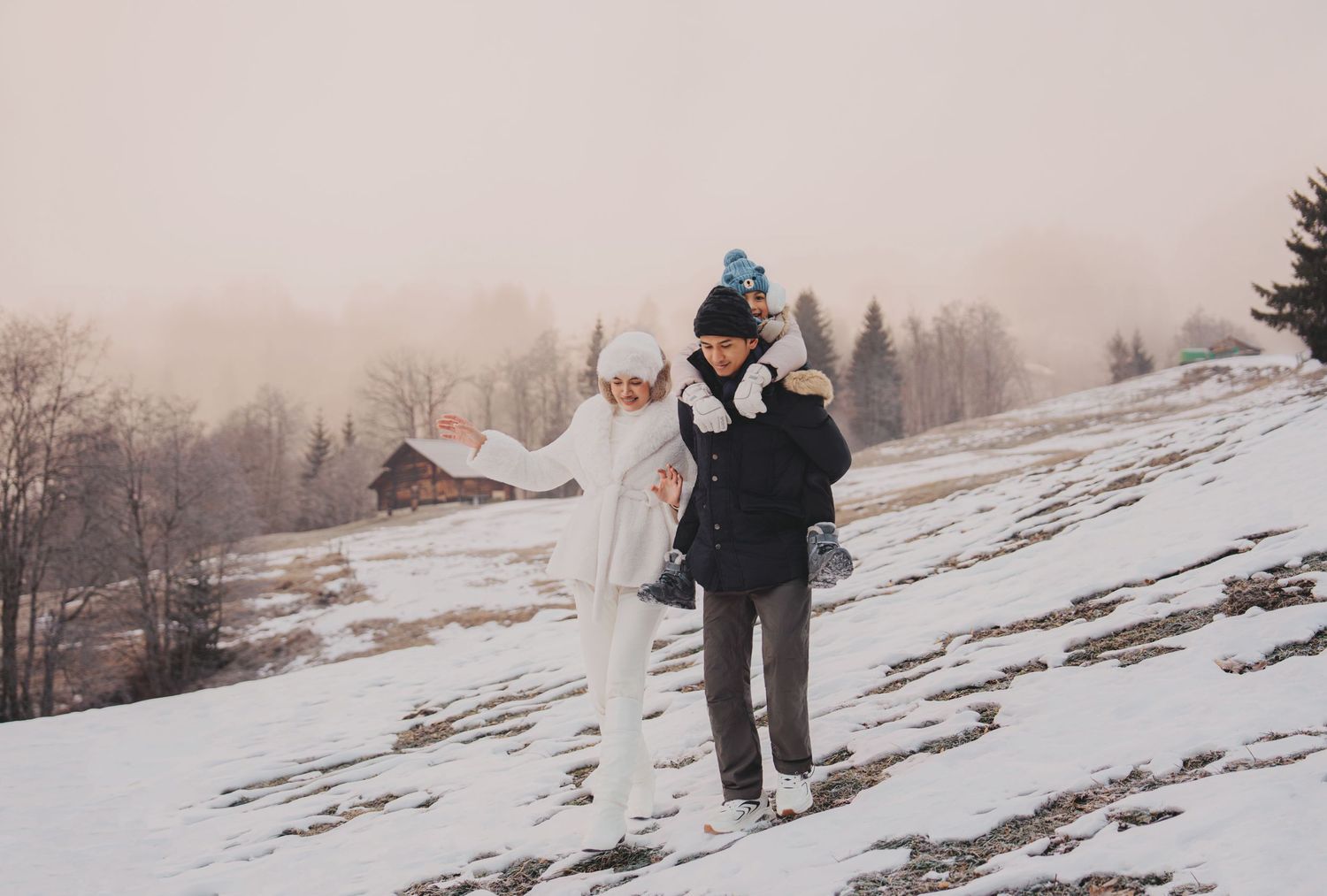 Family walking and playing on snowy hillside with misty winter landscape and bare trees in background.