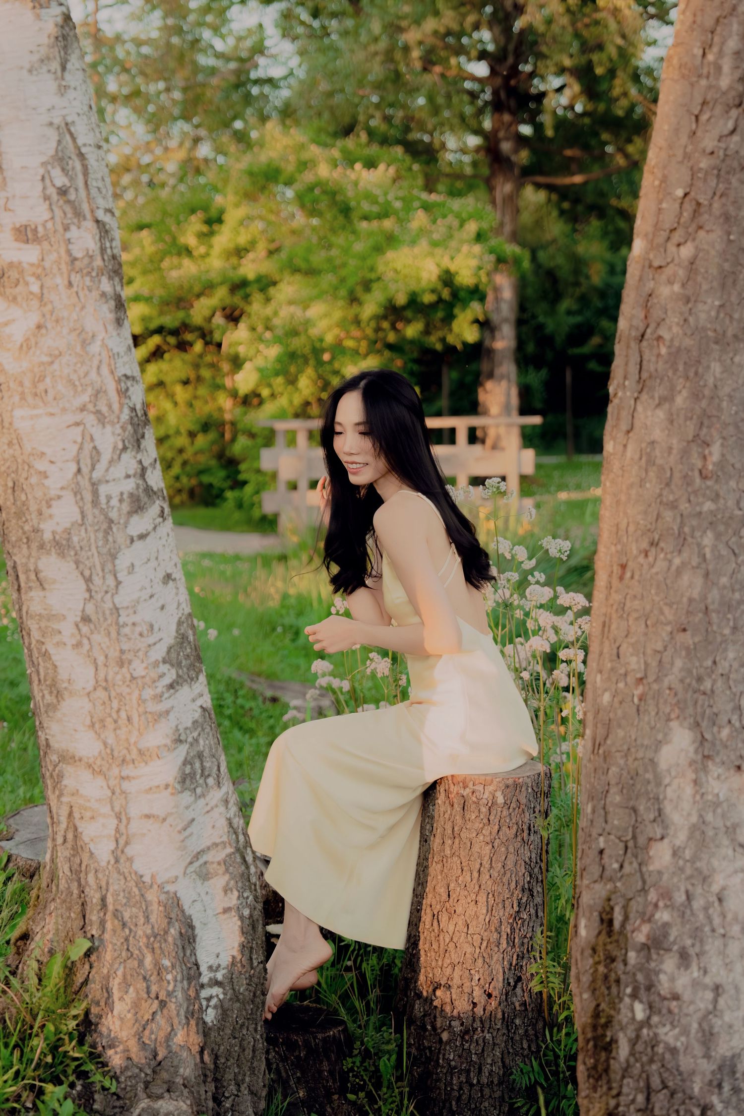 Smiling woman in yellow dress sitting on tree stump in park.