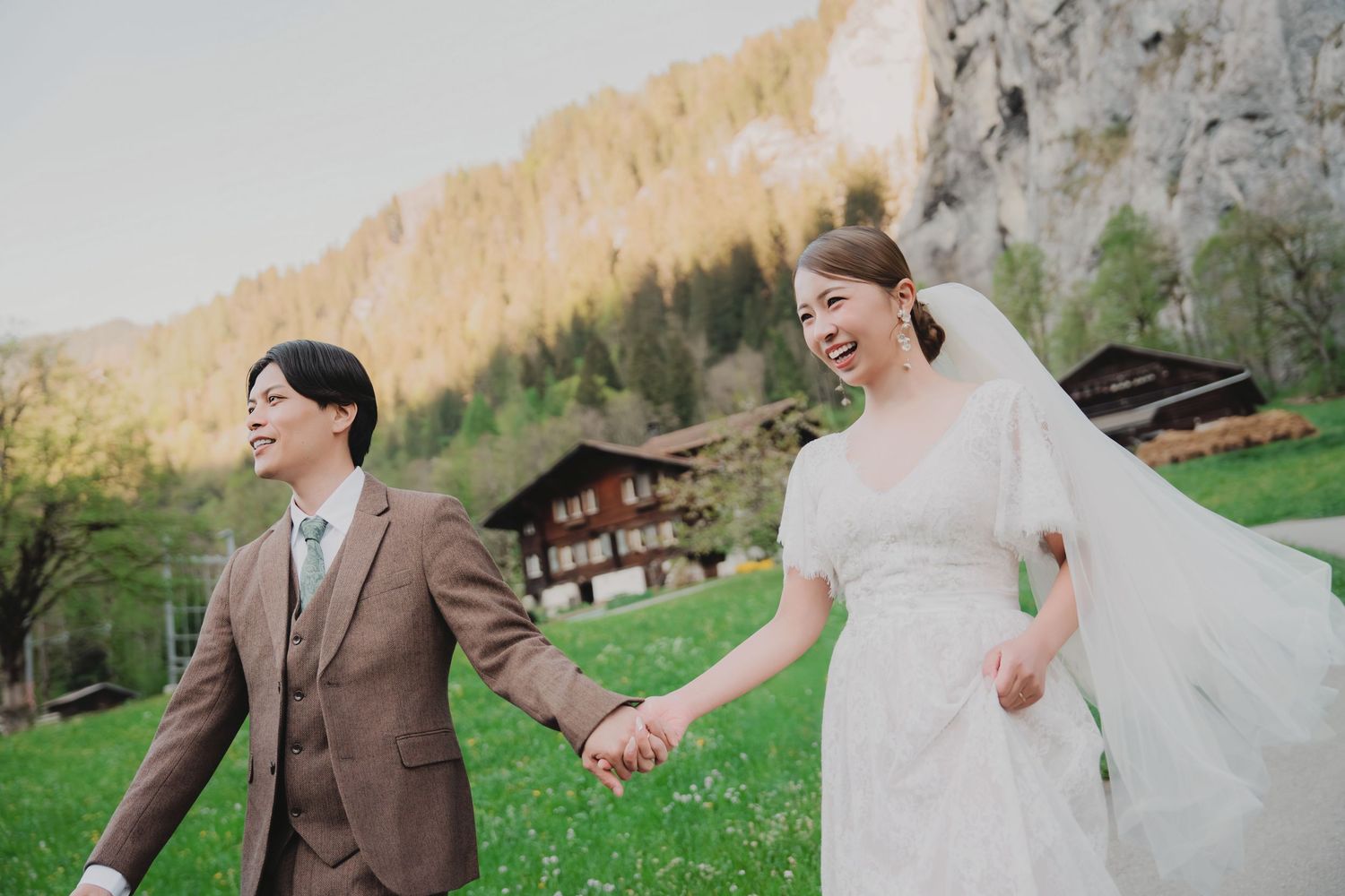 Joyful couple holding hands walking through green meadow with dramatic Swiss mountain cliffs.