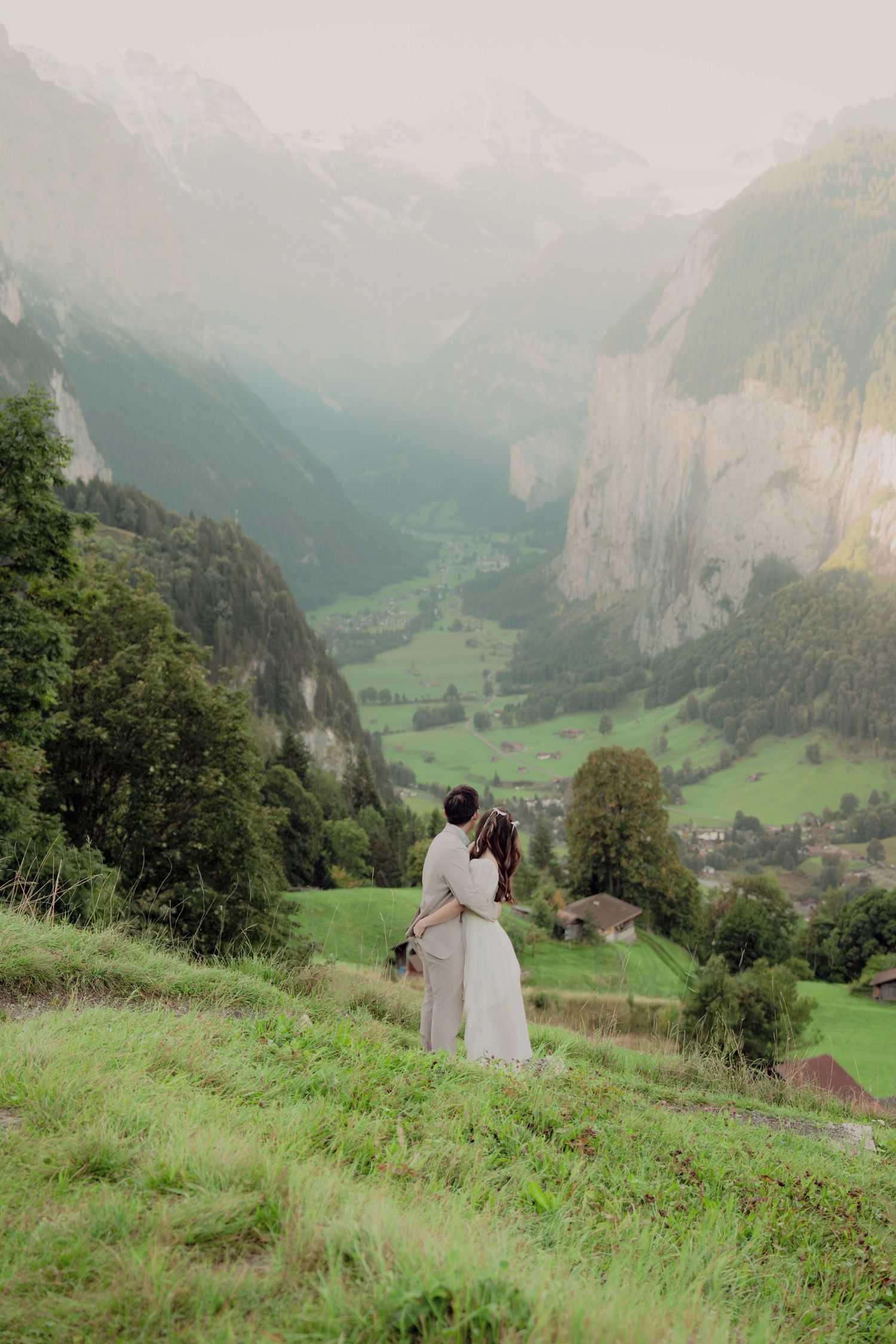 Couple sharing a romantic embrace on a green Swiss hillside with a misty mountain valley stretching below them.