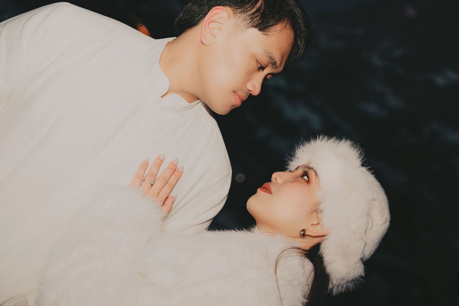 Couple sharing a romantic intimate moment dressed in white winter outfits against a dark background.