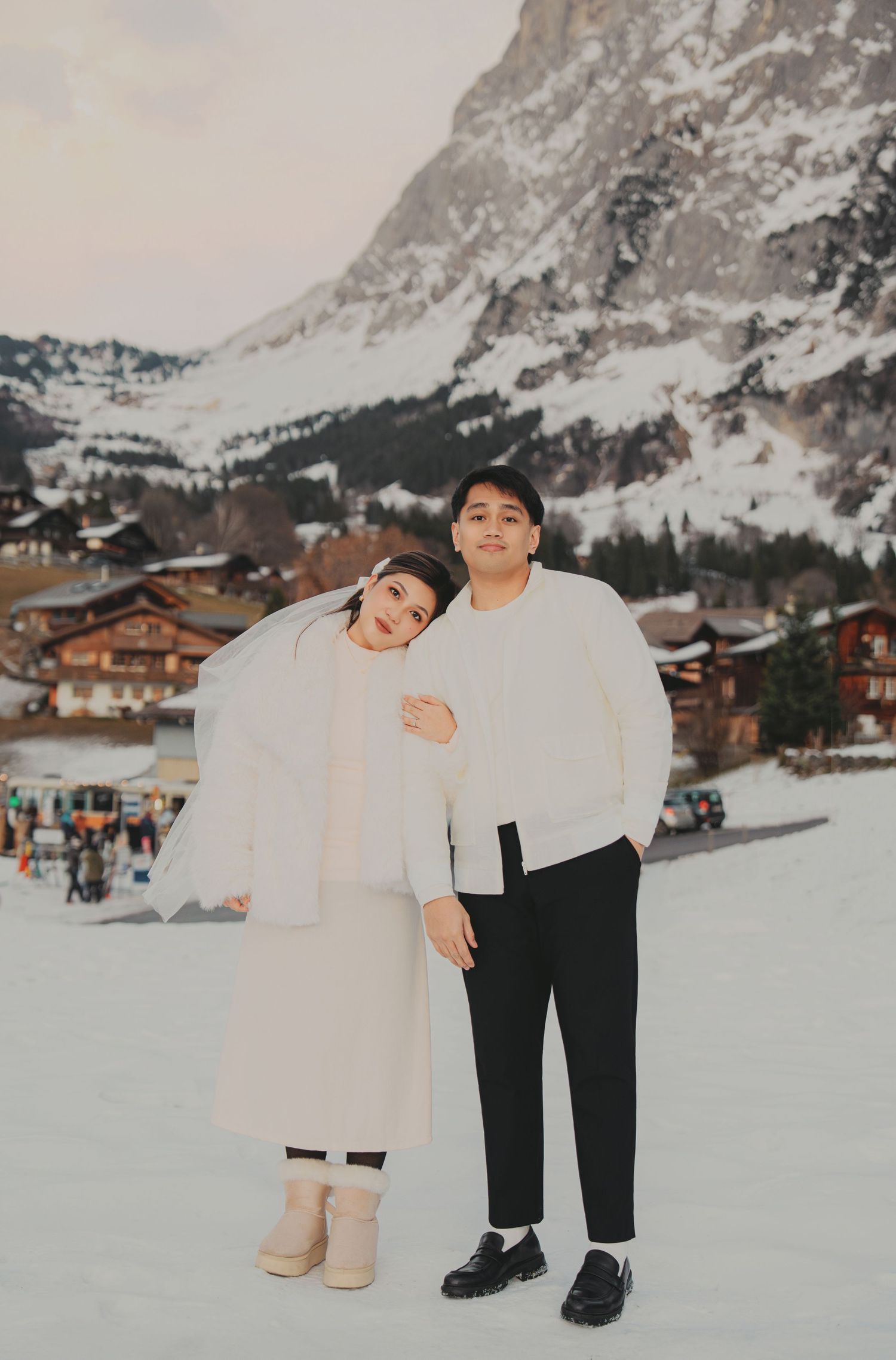 Couple posing together in white winter outfits against a snowy alpine village and mountain backdrop.