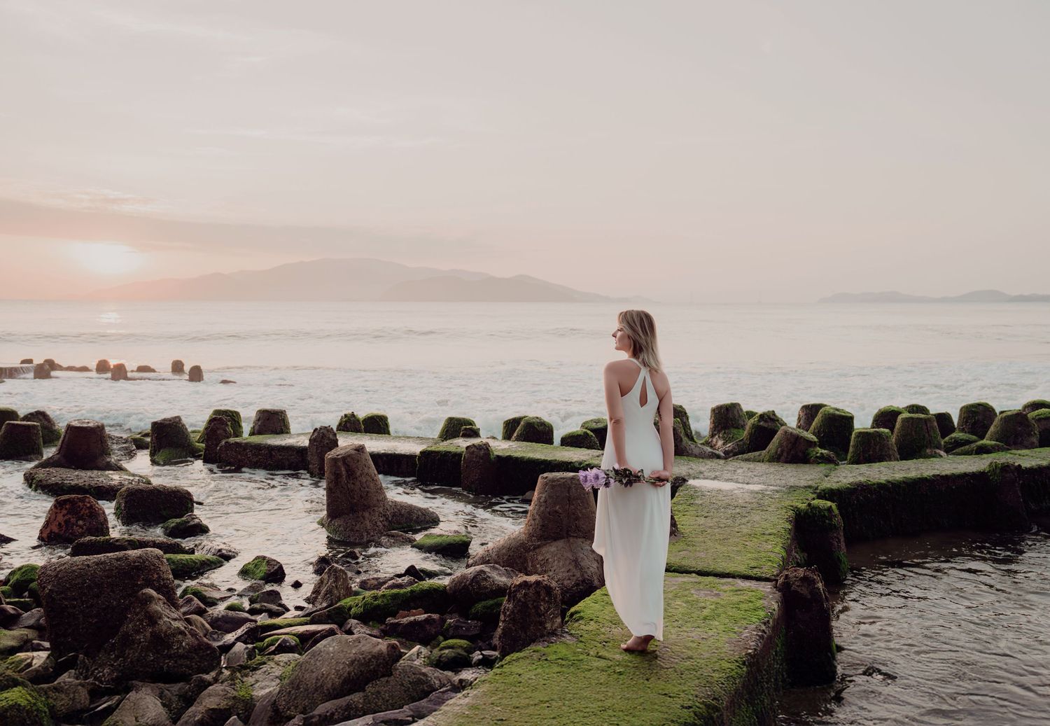 Woman in white dress standing on mossy rocks by the ocean at sunset with mountains in the background.